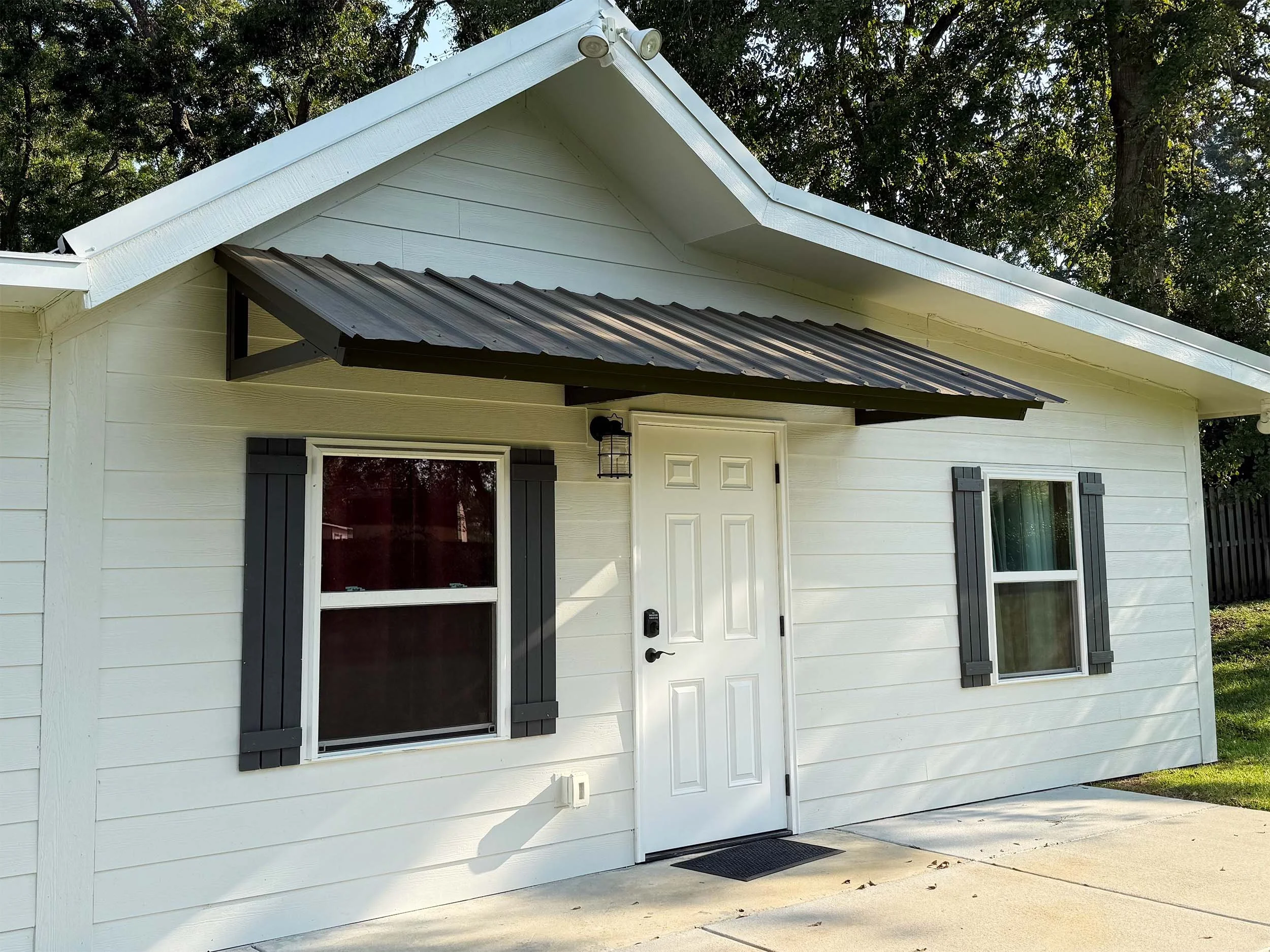 A white house with a door, two windows with black shutters, and a metal awning over the door. The house has a gable roof with white trim, and trees are visible in the background.