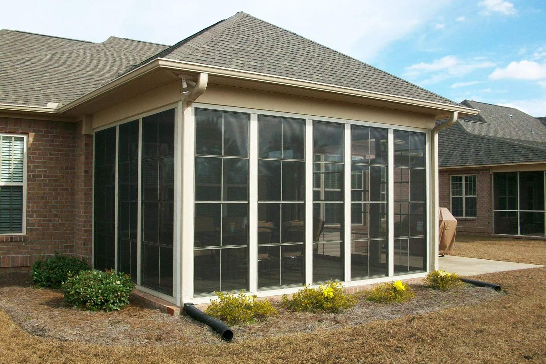 A backyard view of a brick house with a screened-in porch and a sloped roof, with bushes and a lawn in the foreground and neighboring houses in the background.