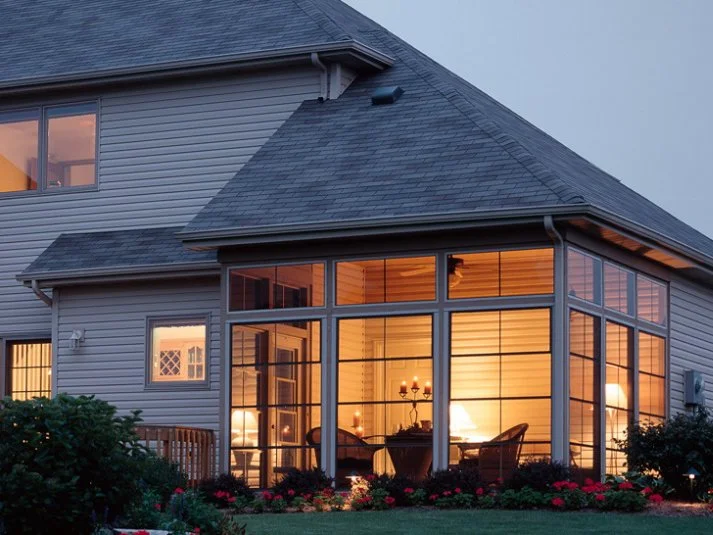The image shows a house with a glass enclosed porch illuminated from within, revealing a dining area with a chandelier and chairs. The house has gray siding and a sloped roof, with gardens and bushes in the foreground.