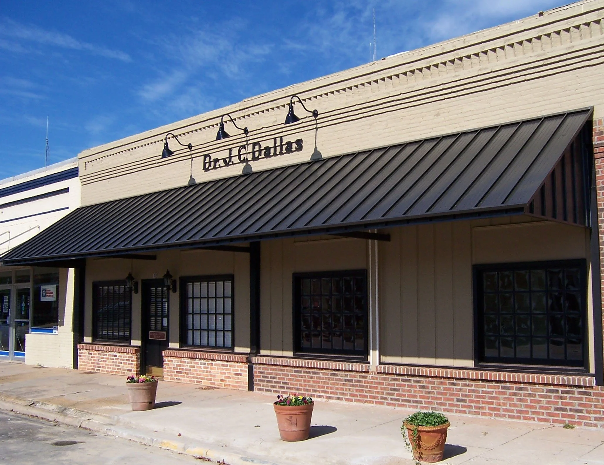 The facade of a commercial building with a sign that reads 'Dr. C. Challas' under three black pendant lights, with large windows, a brick base, and potted plants on the sidewalk, with a bright blue sky overhead.