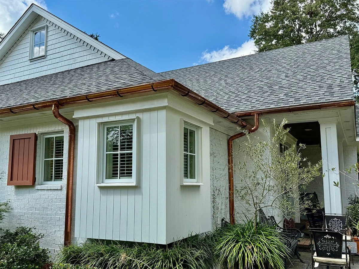 Exterior view of a house with white brick and siding, shingled roof, copper gutters, and a small patio with black metal chairs and greenery.