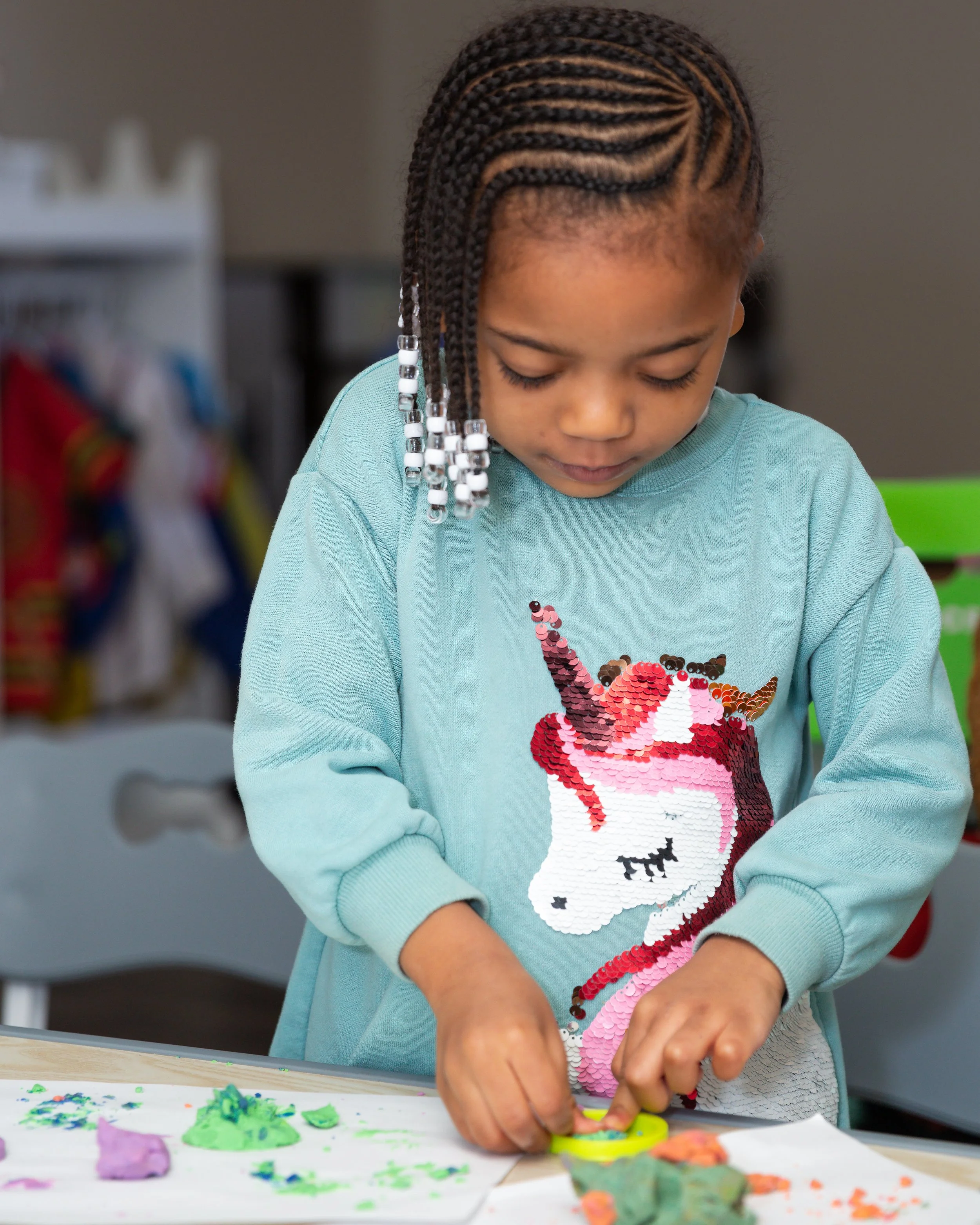 A young girl with braided hair decorated with beads, working on a craft project at a table with colorful modeling clay and tools.