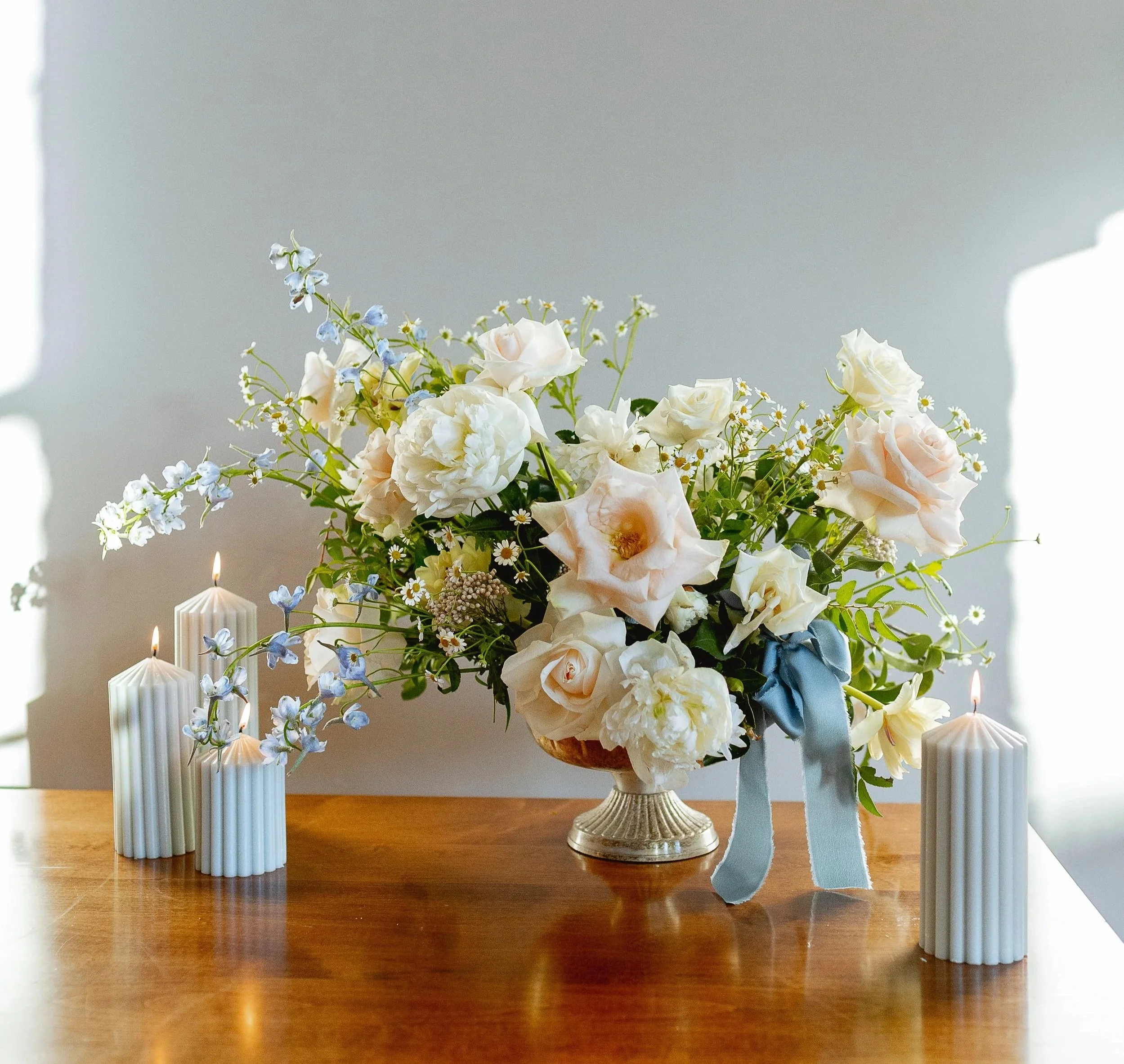 A floral arrangement with white and pale pink flowers in a metallic vase, decorated with a blue ribbon, on a wooden table, flanked by four white pillar candles.