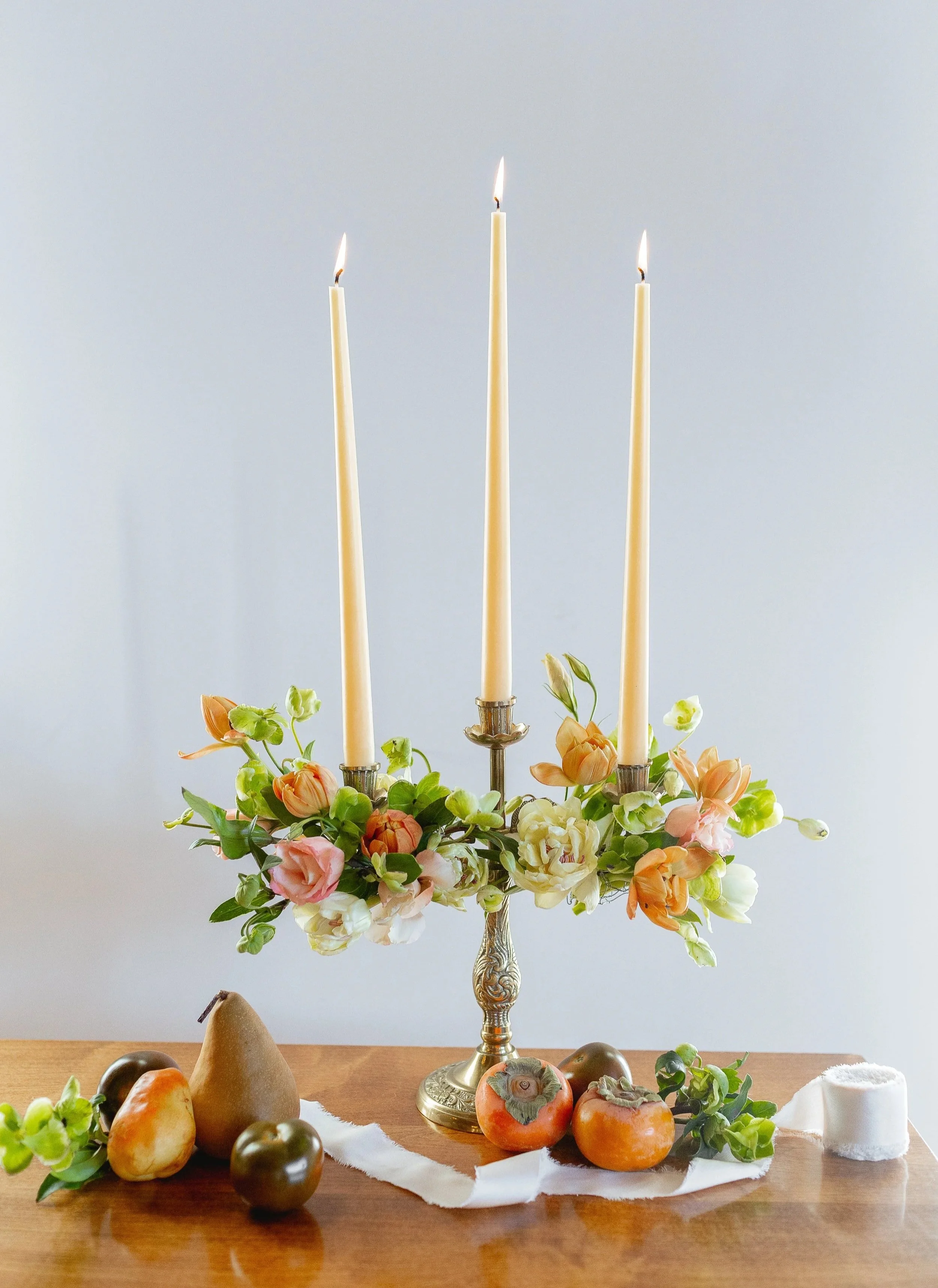 A decorative table arrangement with a silver candelabrum holding three lit candles, surrounded by peach, white, and orange flowers and various fruits on a white cloth on a wooden table.