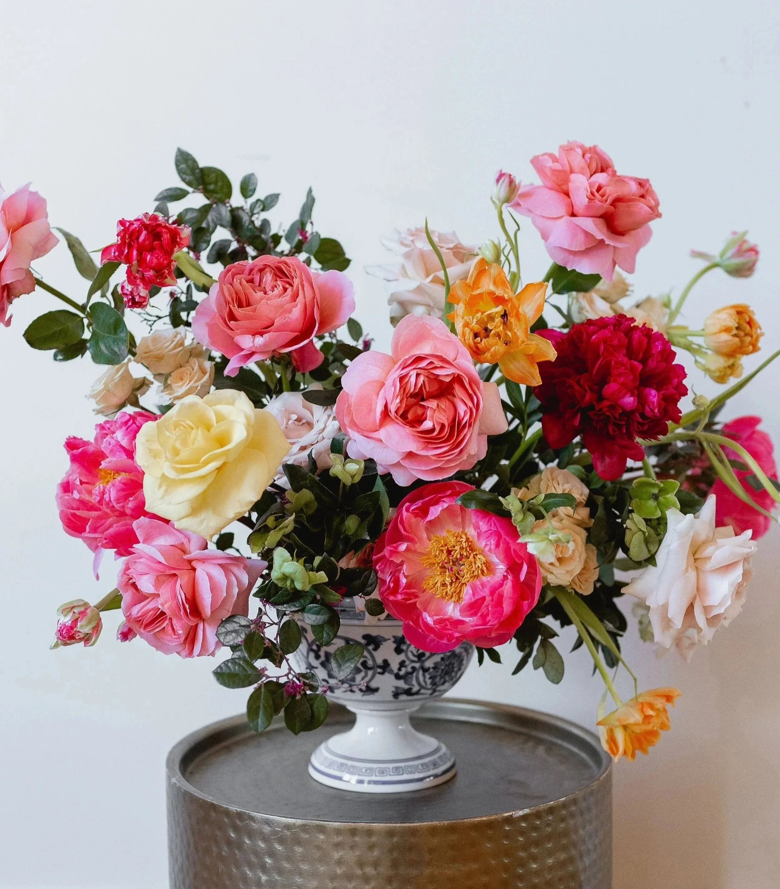 A bouquet of pink, yellow, red, and orange roses in a decorative white and blue vase on a round metallic table against a plain white wall.