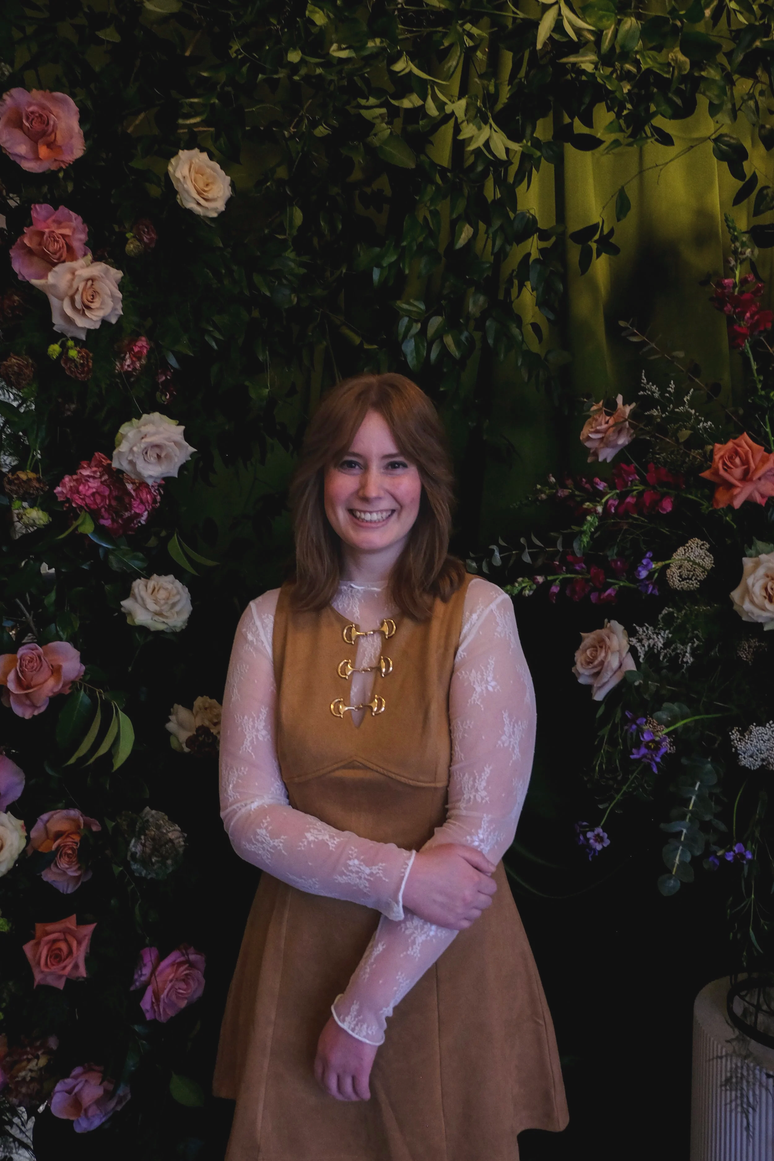 A smiling woman with shoulder-length brown hair stands in front of a backdrop covered with colorful flowers and green foliage.