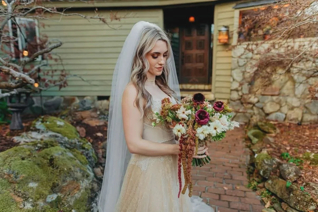A bride in a light-colored dress with a veil holding a bouquet of flowers, standing outdoors in front of a house with stone and wood exterior, surrounded by rocks and autumn foliage.