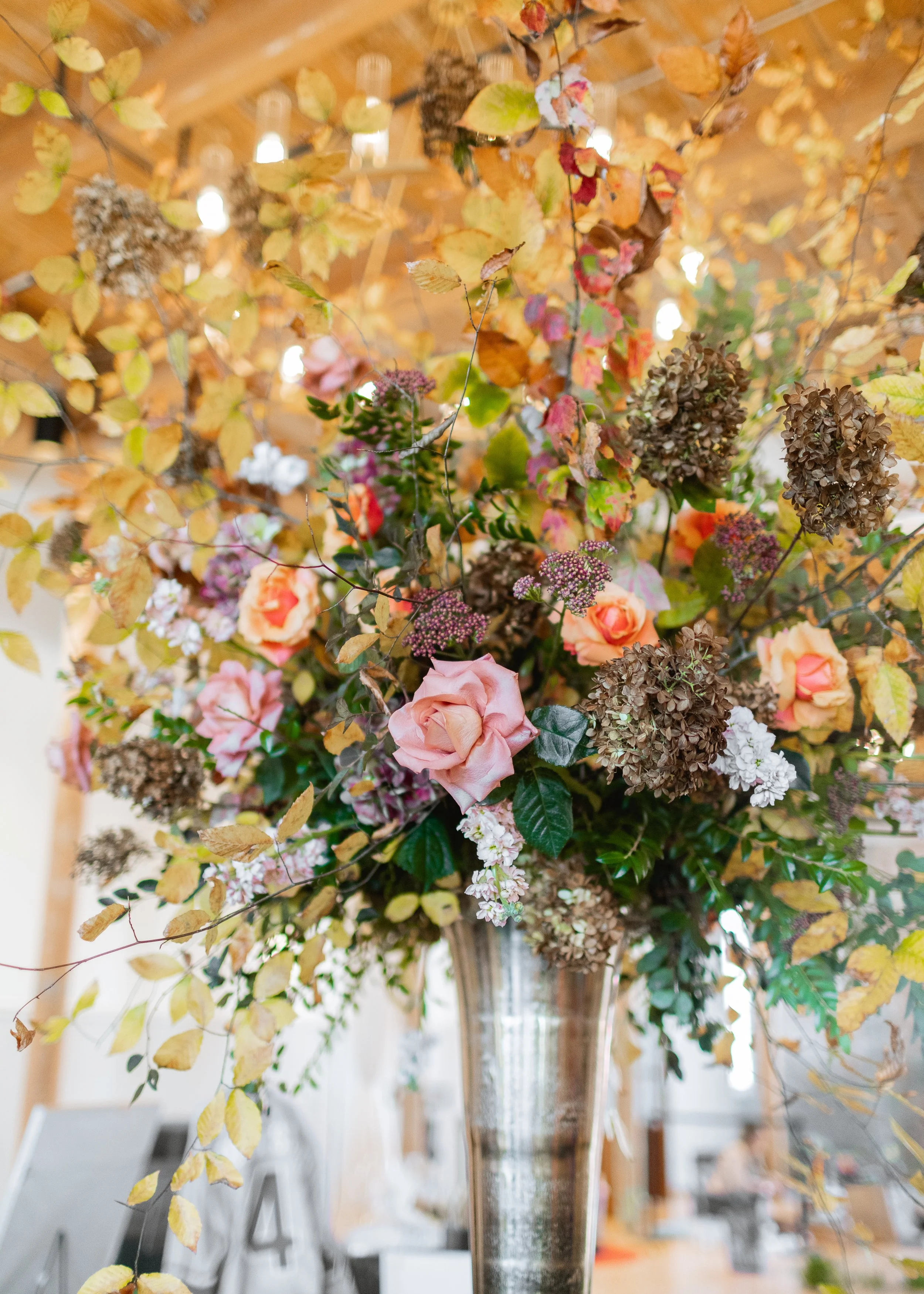 A large bouquet of autumn-colored flowers and foliage in a tall, shiny metallic vase, set on a table in what appears to be a decorated indoor setting.