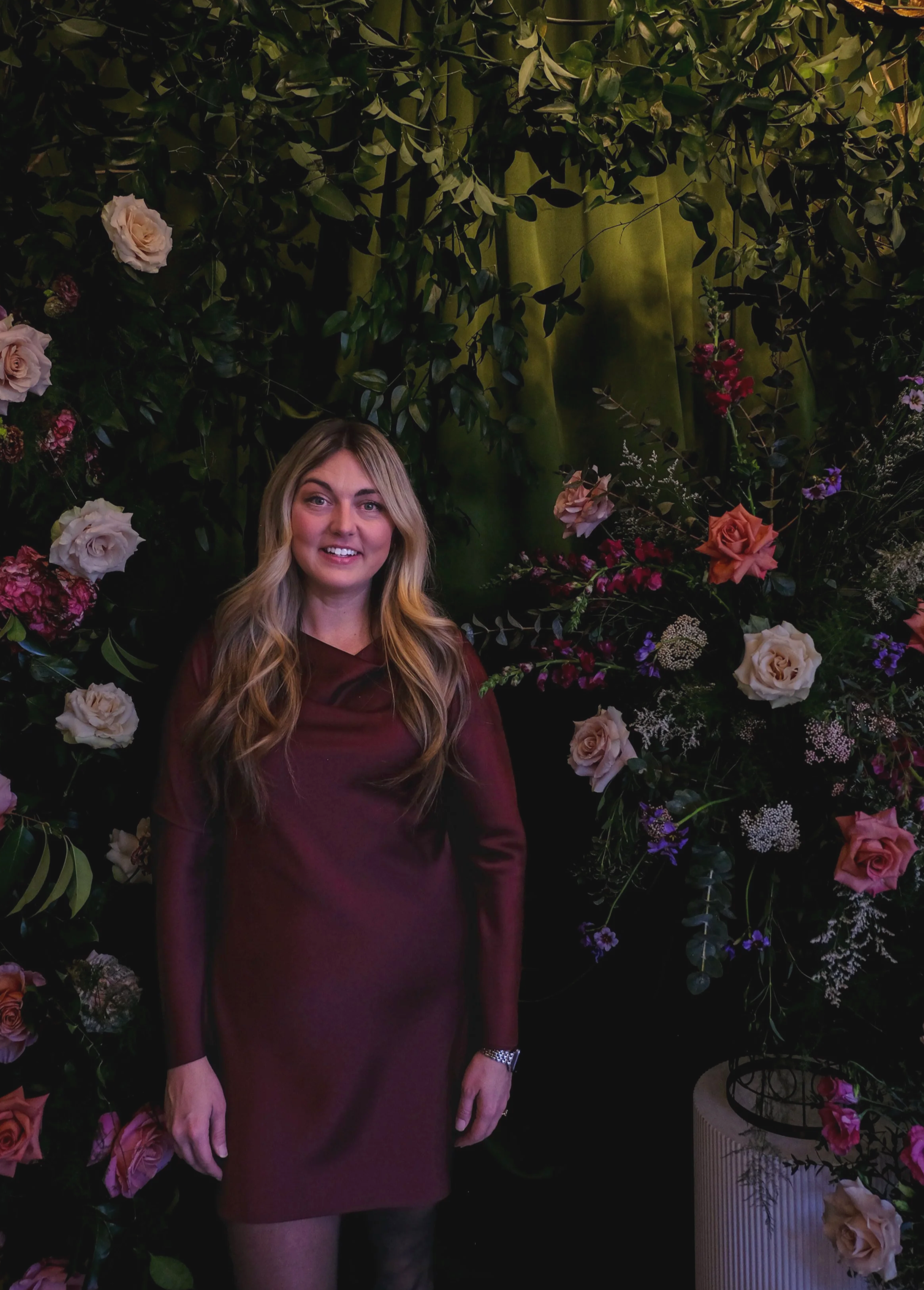 A woman with long wavy blonde hair wearing a dark red dress with long sleeves, standing in front of a floral backdrop with pink, peach, and purple flowers and green foliage.