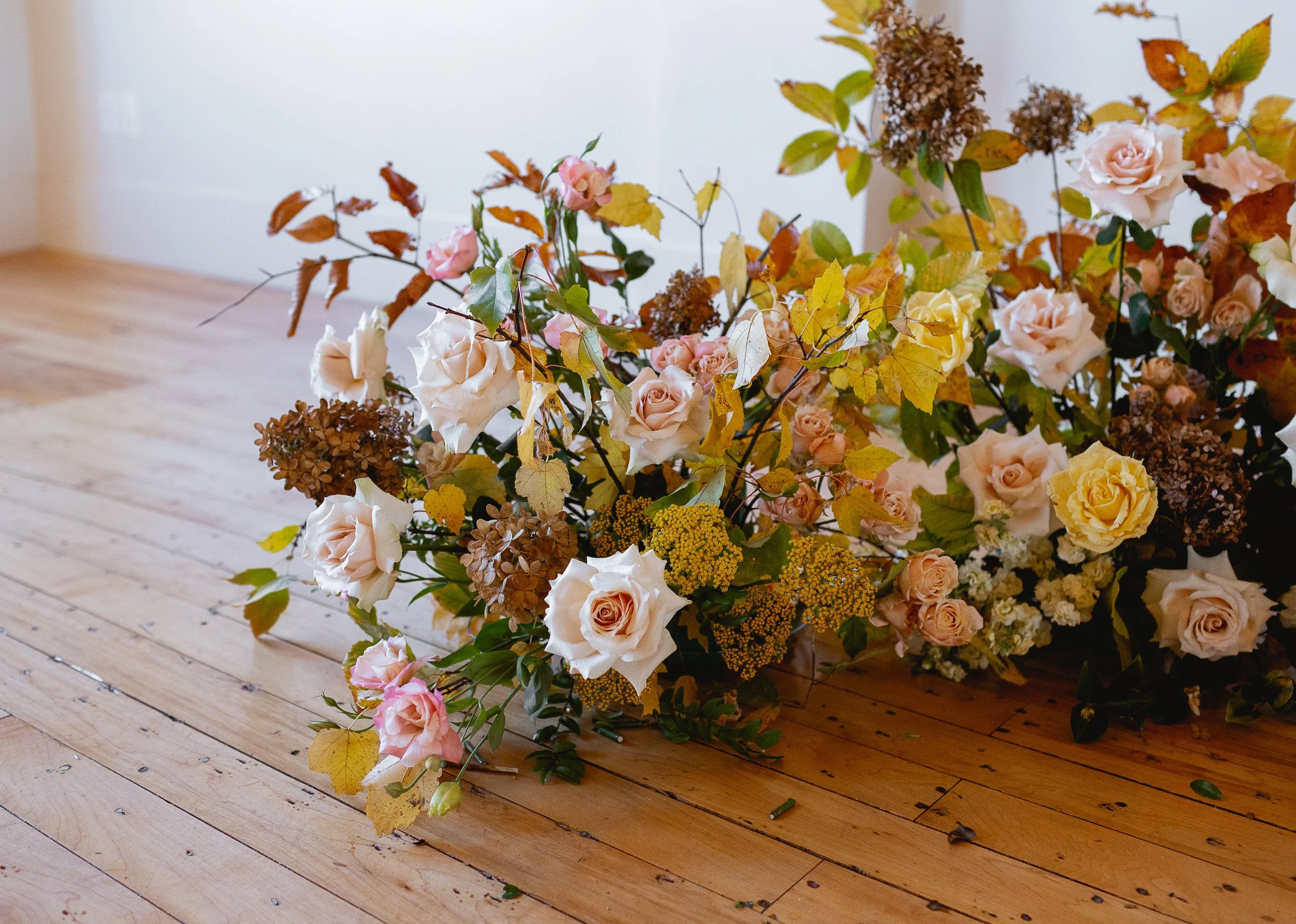 A large arrangement of roses, hydrangeas, and other flowers in various shades of pink, yellow, and brown lying on a wooden floor.