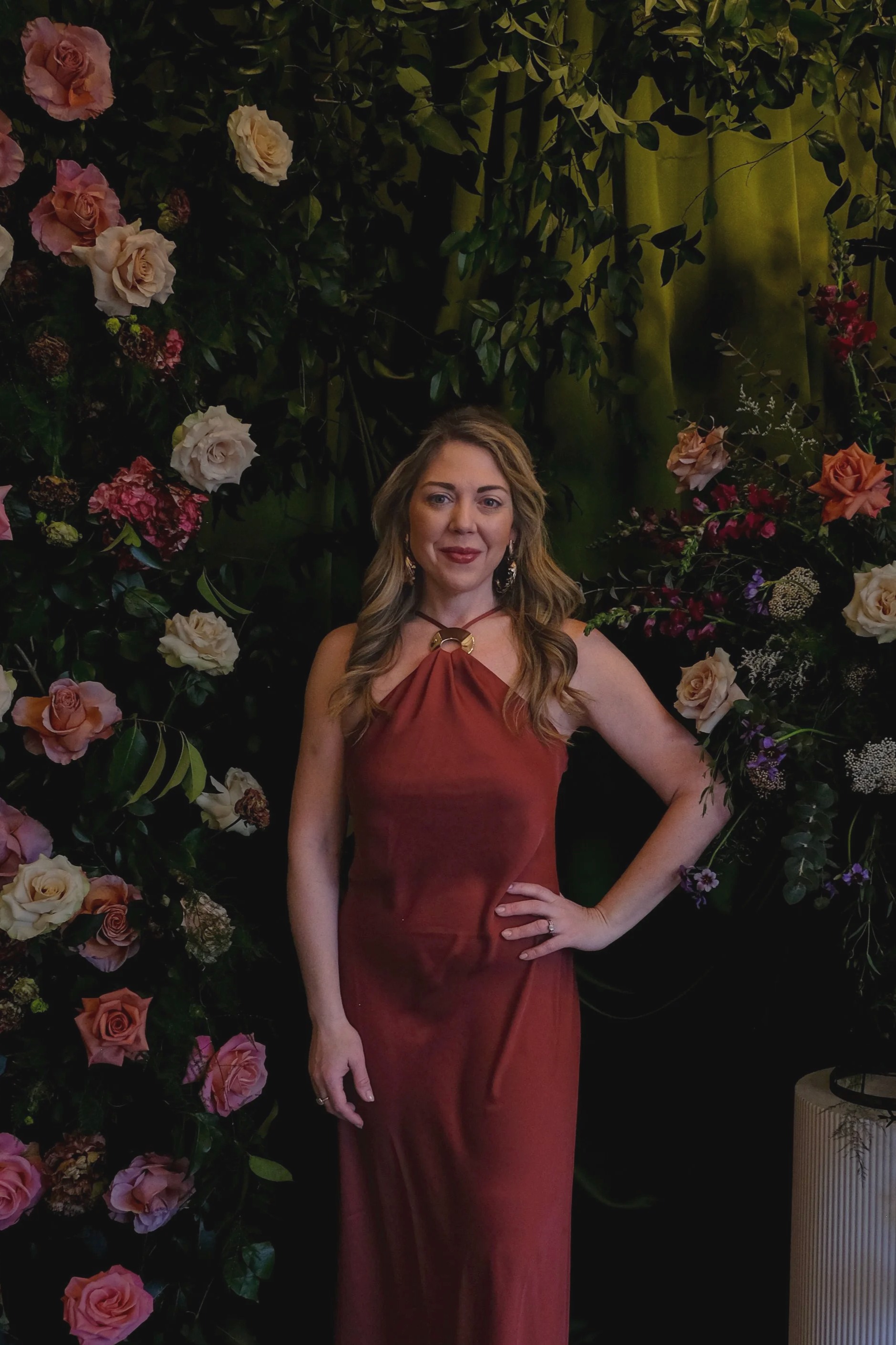 A woman in a sleeveless, reddish-brown dress standing in front of a backdrop of pink, peach, and cream roses and greenery, indoors with dark and light green curtains in the background.