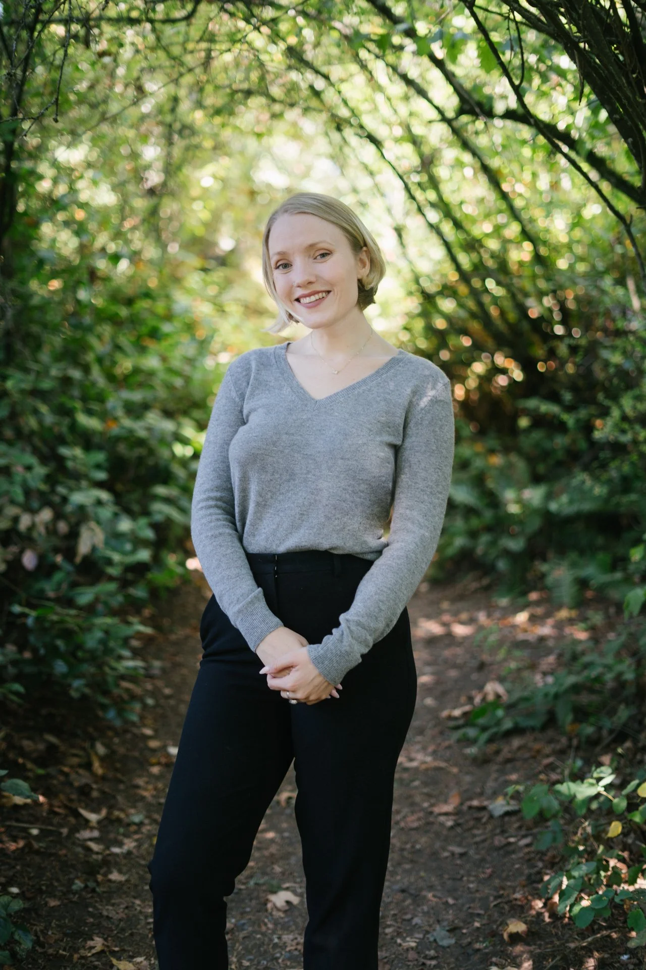 A smiling woman with short blonde hair standing outdoors on a nature trail surrounded by green foliage and trees.