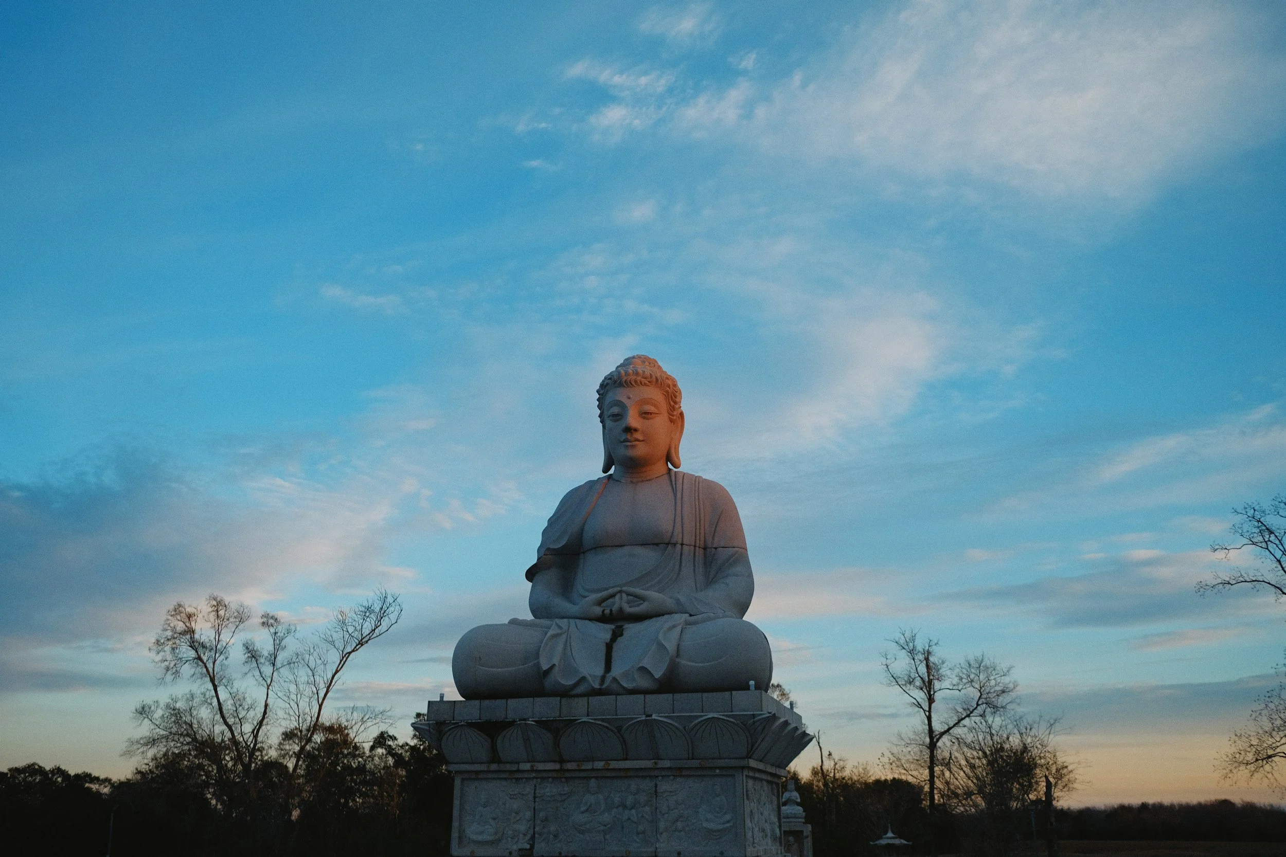 A large white Buddha statue sitting in a meditative pose outdoors under a blue sky with some clouds and trees around.