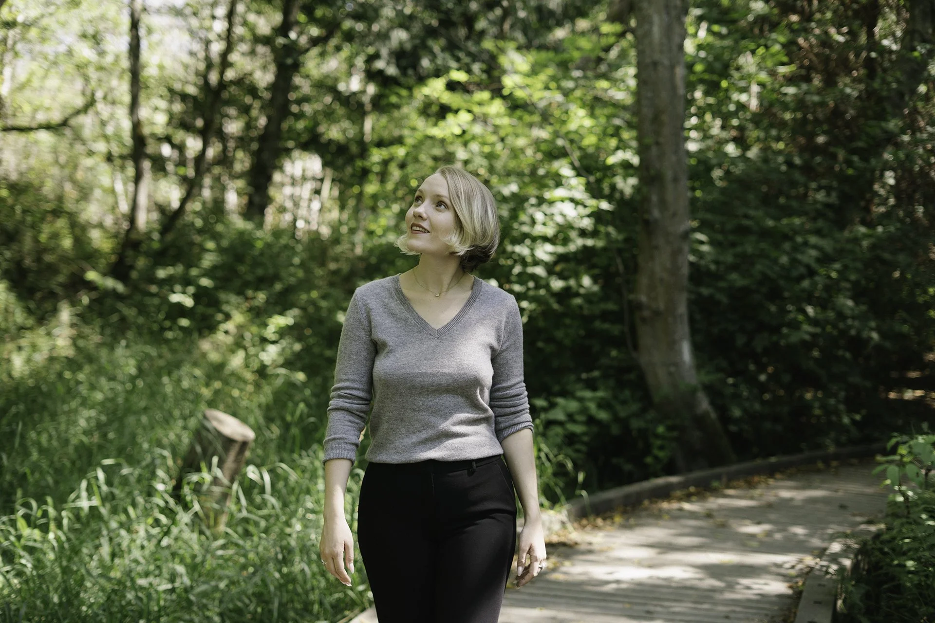 A woman with short blonde hair wearing a gray long-sleeve shirt and black pants walking along a wooded path.