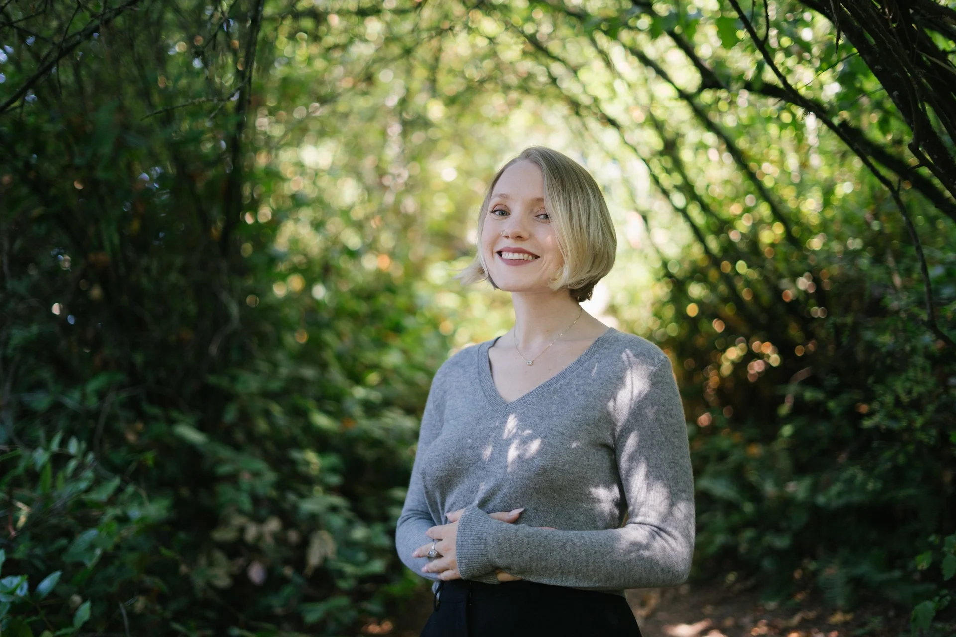A woman with shoulder-length blonde hair smiling outdoors in front of green foliage and sunlight filtering through the trees.
