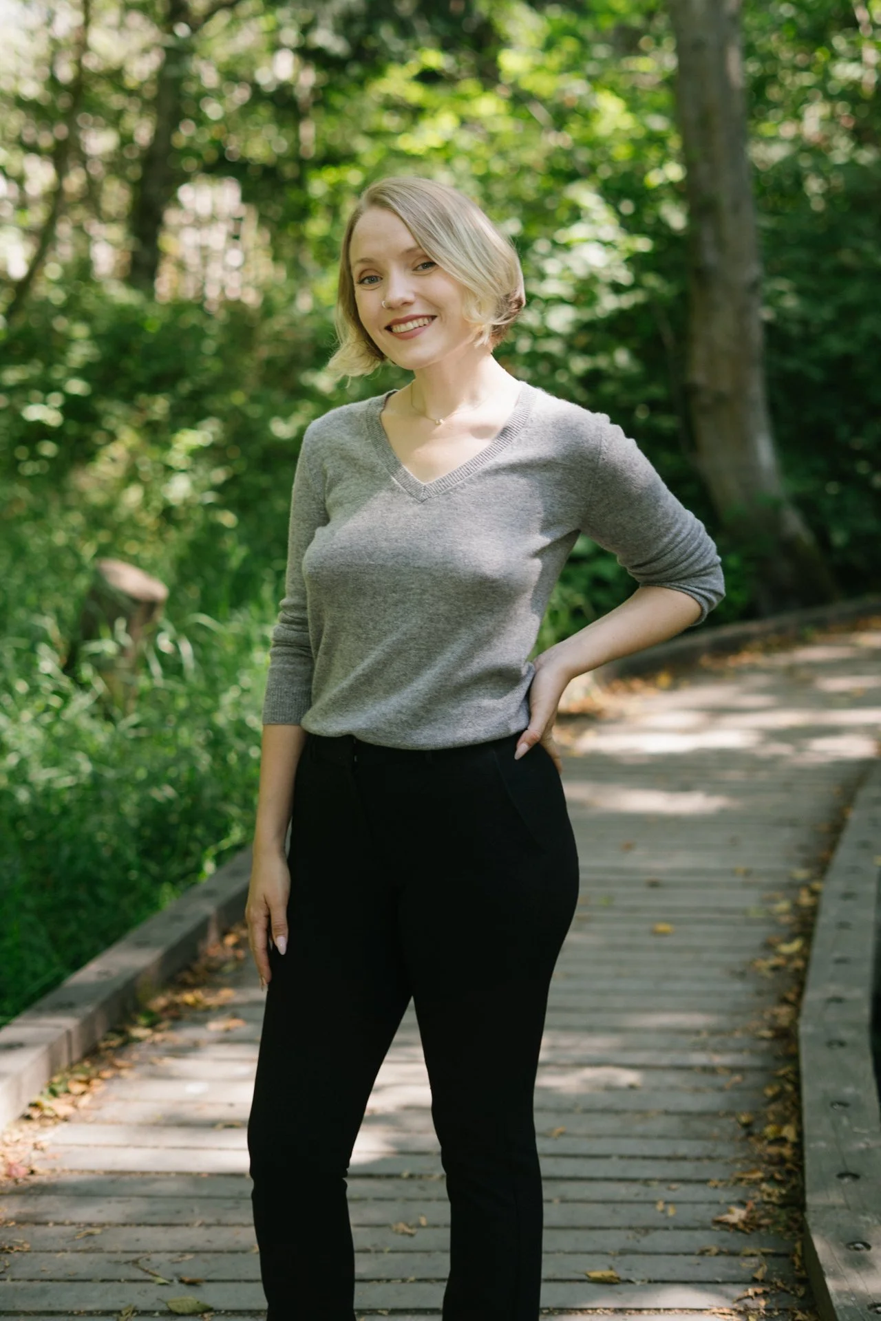 A young woman with short blonde hair smiling outdoors on a wooden pathway surrounded by green trees and foliage.
