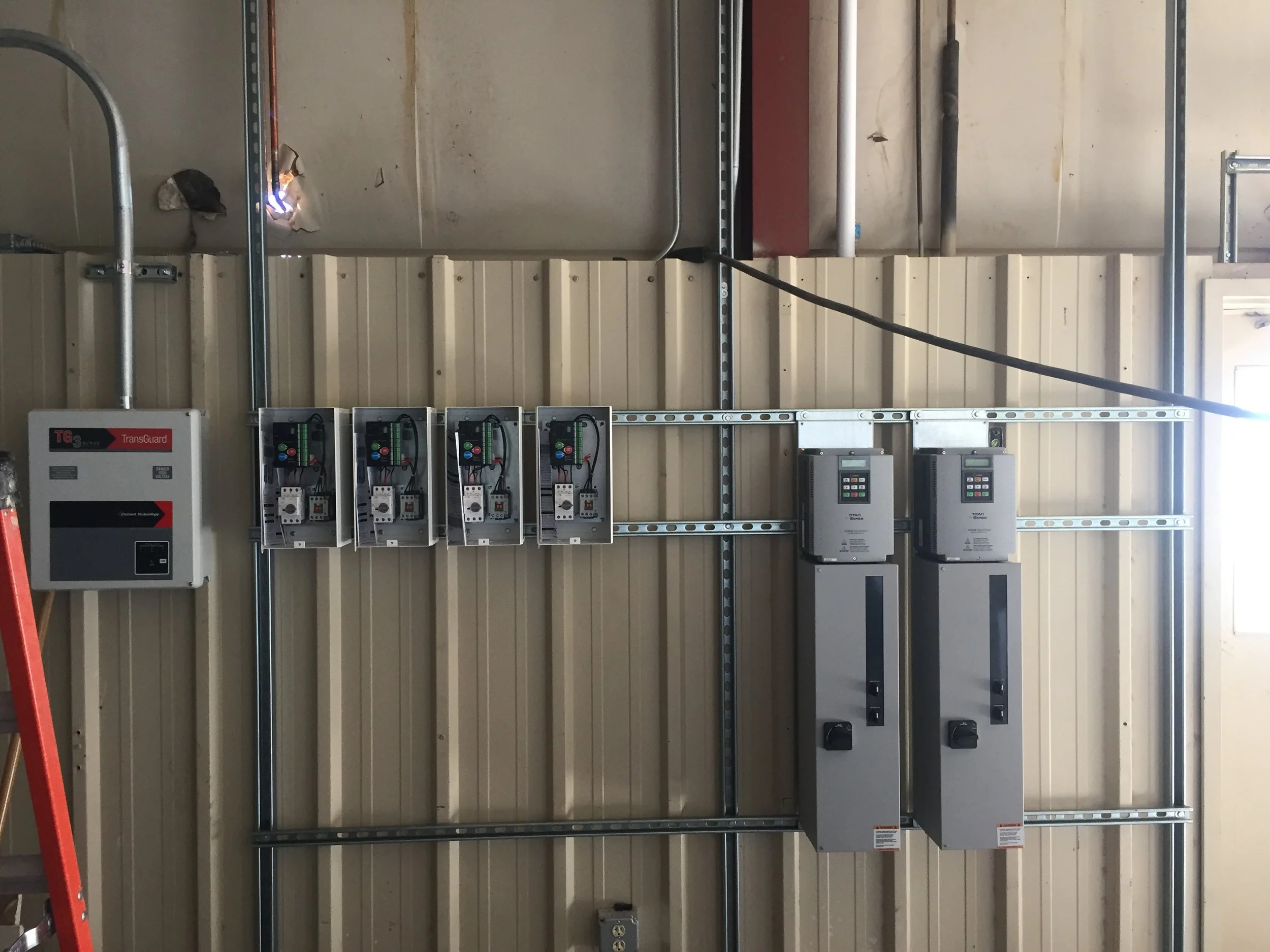 Electrical control panels, circuit breakers, and wiring mounted on a metal grid wall inside a utility room or industrial setting.
