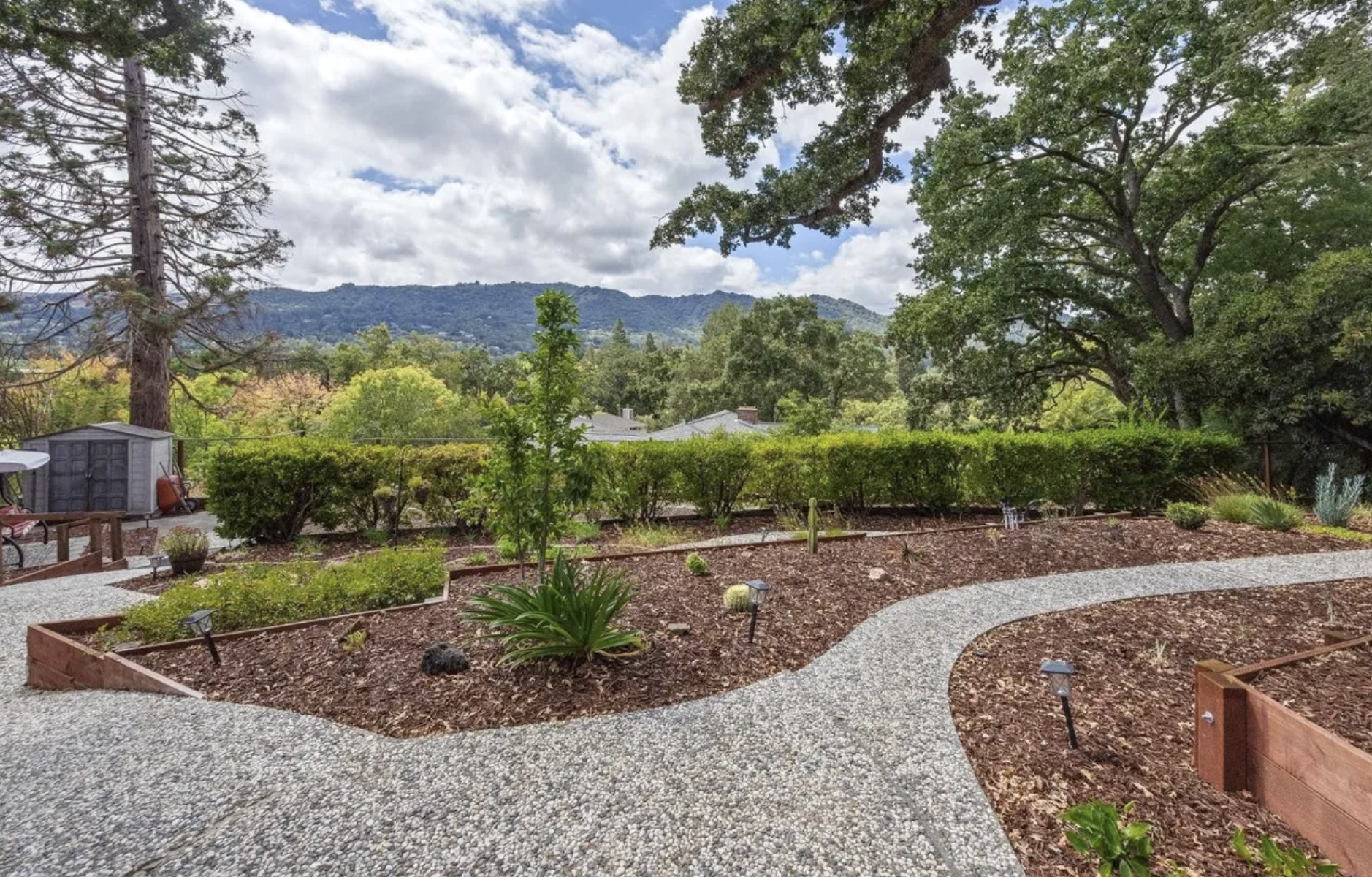 A landscaped yard with a gravel pathway winding through garden beds, surrounded by green trees and bushes, with hills and a cloudy sky in the background.