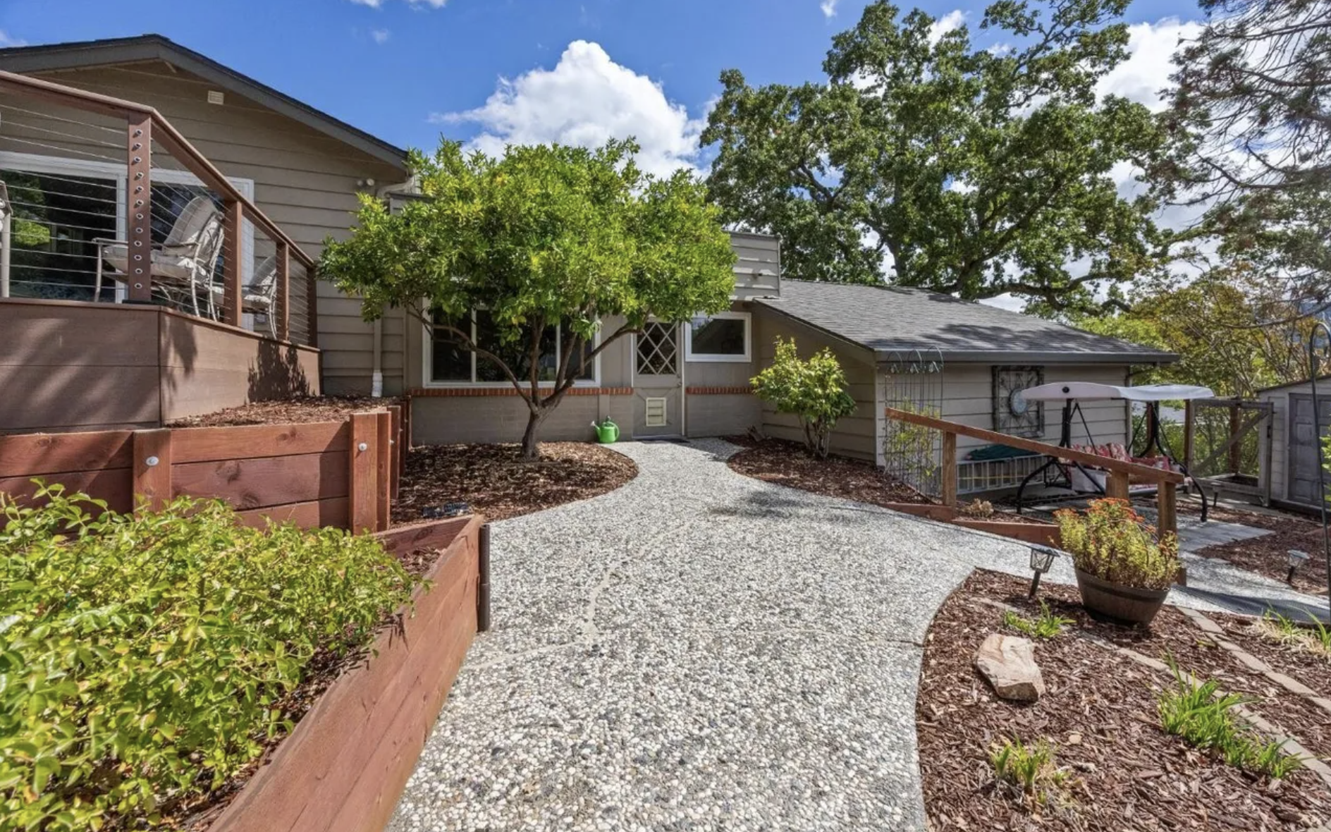 A backyard with a curved gravel pathway, wooden fencing, trees, potted plants, a patio with outdoor furniture, and a house with grey siding