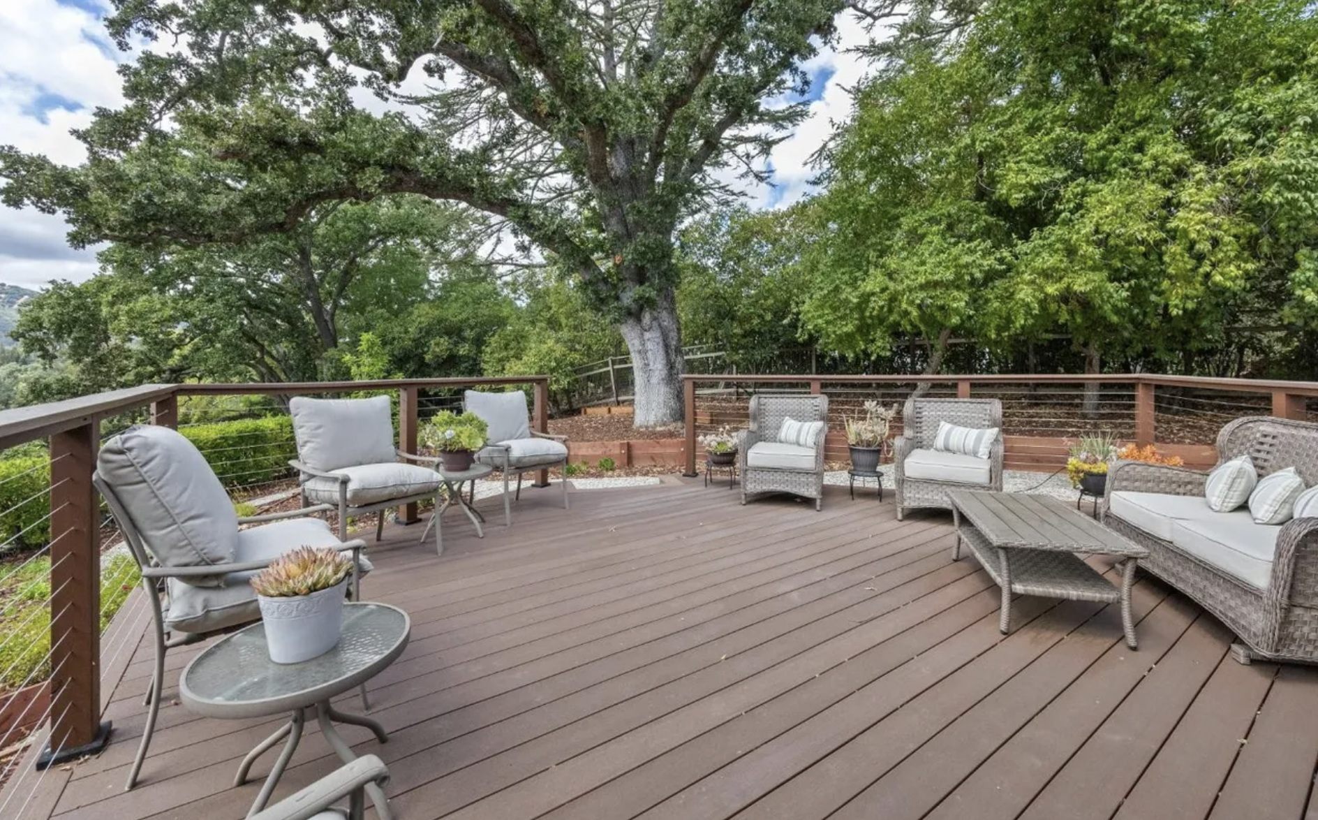 Outdoor wooden deck with seating and potted plants, surrounded by green trees and a partly cloudy sky.