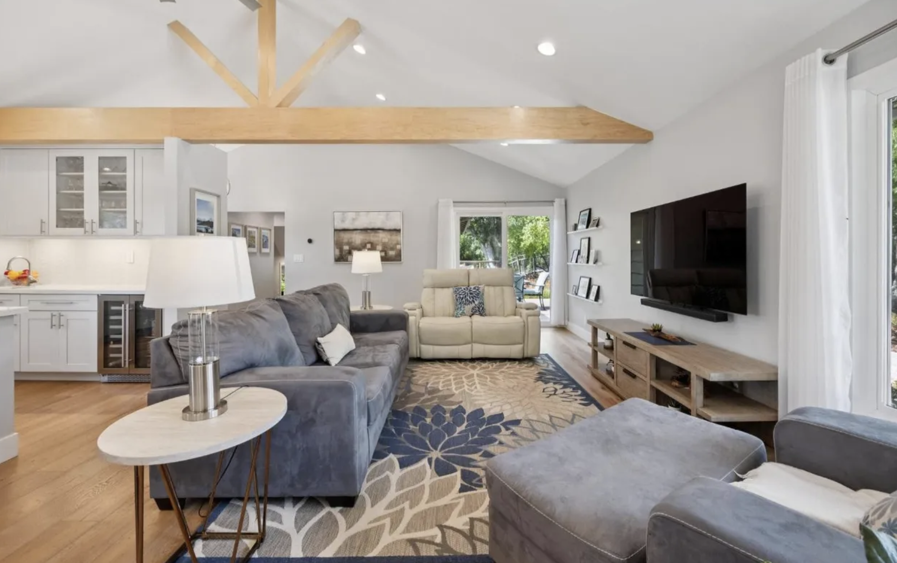 Living room with gray sofas, a white reclining chair, a wooden TV stand with a large flat-screen TV, a patterned rug, white curtains, and a sliding glass door leading outside.