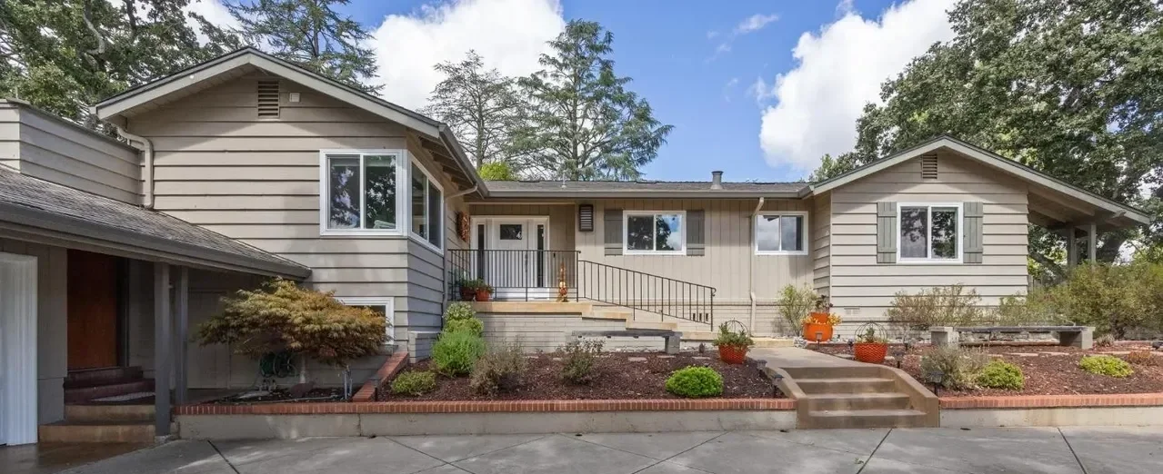 Front view of a beige, split-level house with a landscaped yard, steps leading to the porch, and trees in the background.