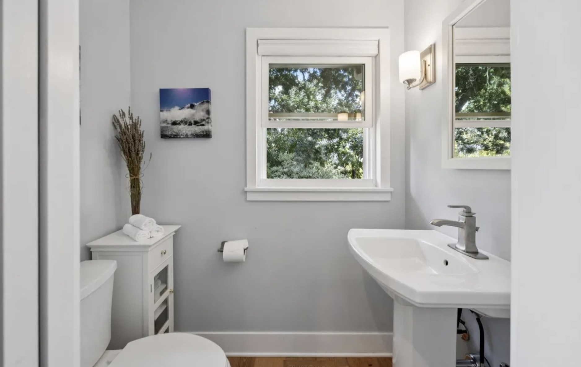 A small, bright bathroom with two windows showing green trees outside. It has a white sink, a mirror, a wall-mounted light, a small cabinet with towels, a picture of ocean waves, and a bundle of dried plants.