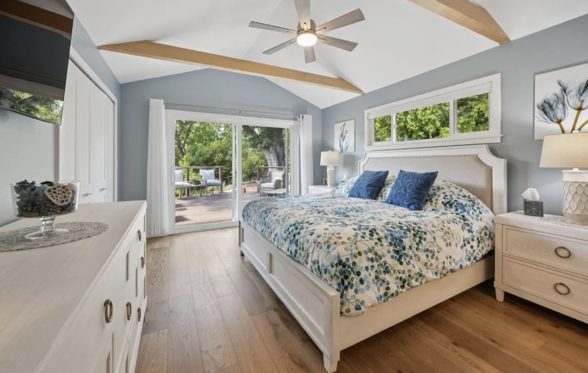 Bright bedroom with a large bed, white furniture, and sliding glass door leading to a balcony with outdoor seating, overlooking greenery.