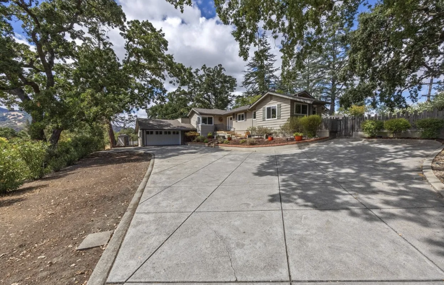 View of a suburban house with a large concrete driveway, trees, and a garden with potted plants, under a partly cloudy sky.