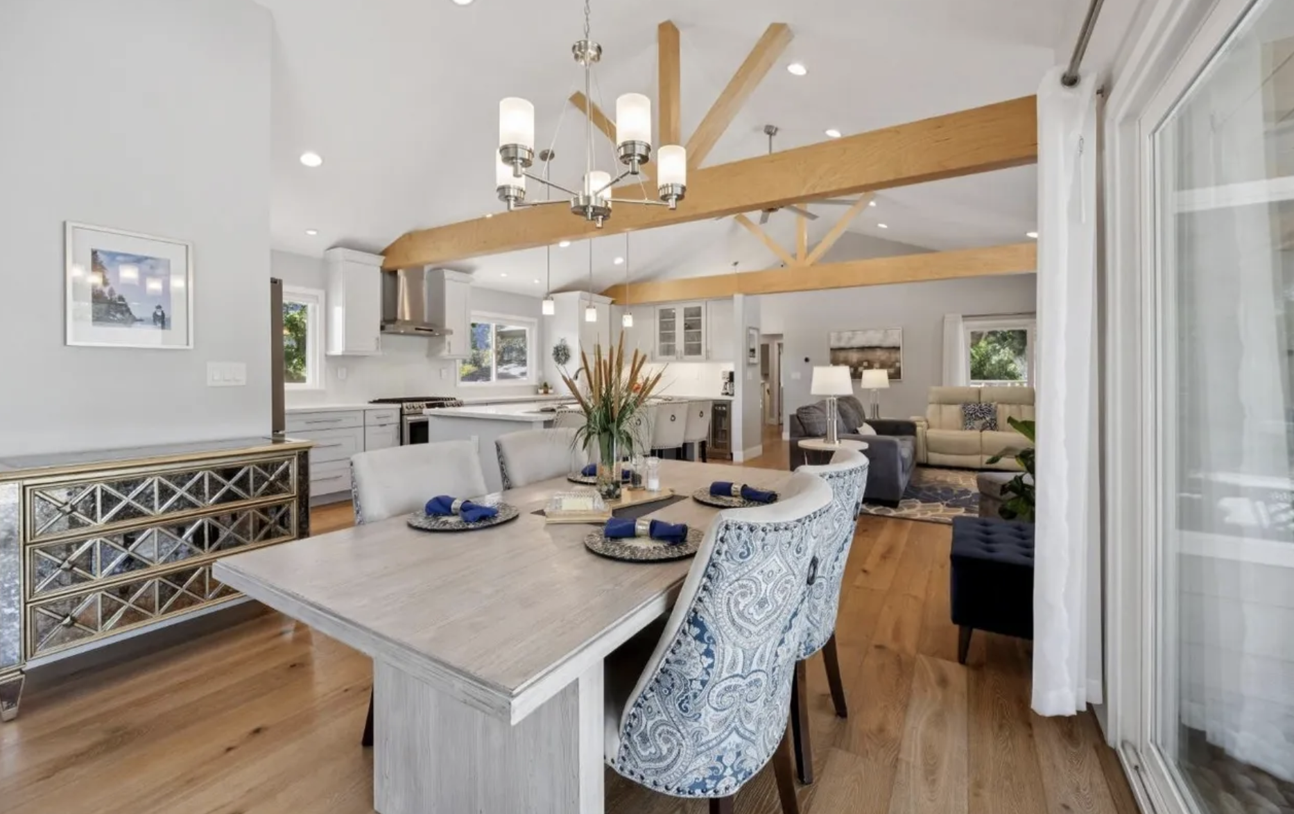 Open-concept dining and living room with white kitchen, wooden beams, and hardwood floors.