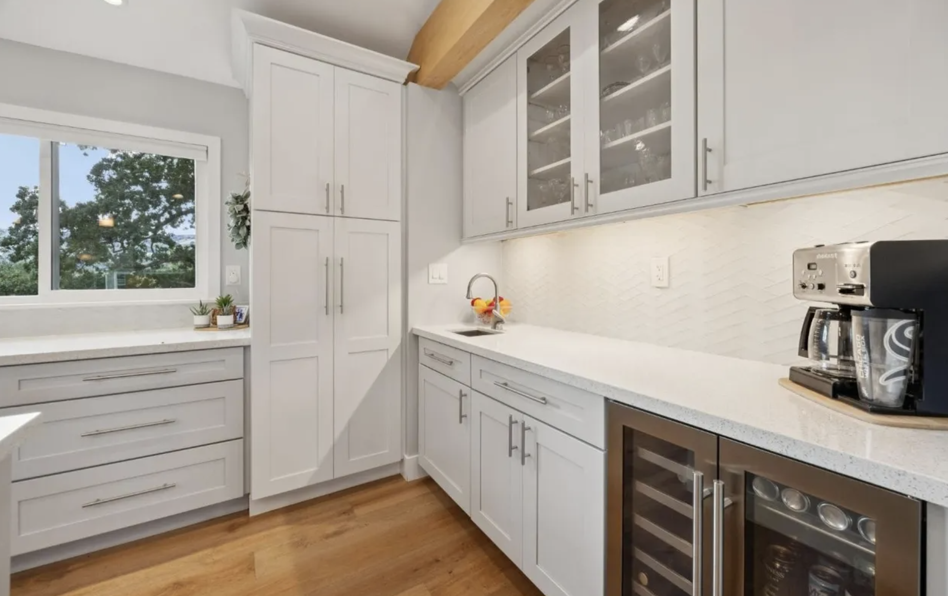 Kitchen with white cabinets, a window, a countertop with a fruit bowl, a coffee maker, and a mini wine cooler, hardwood floors.