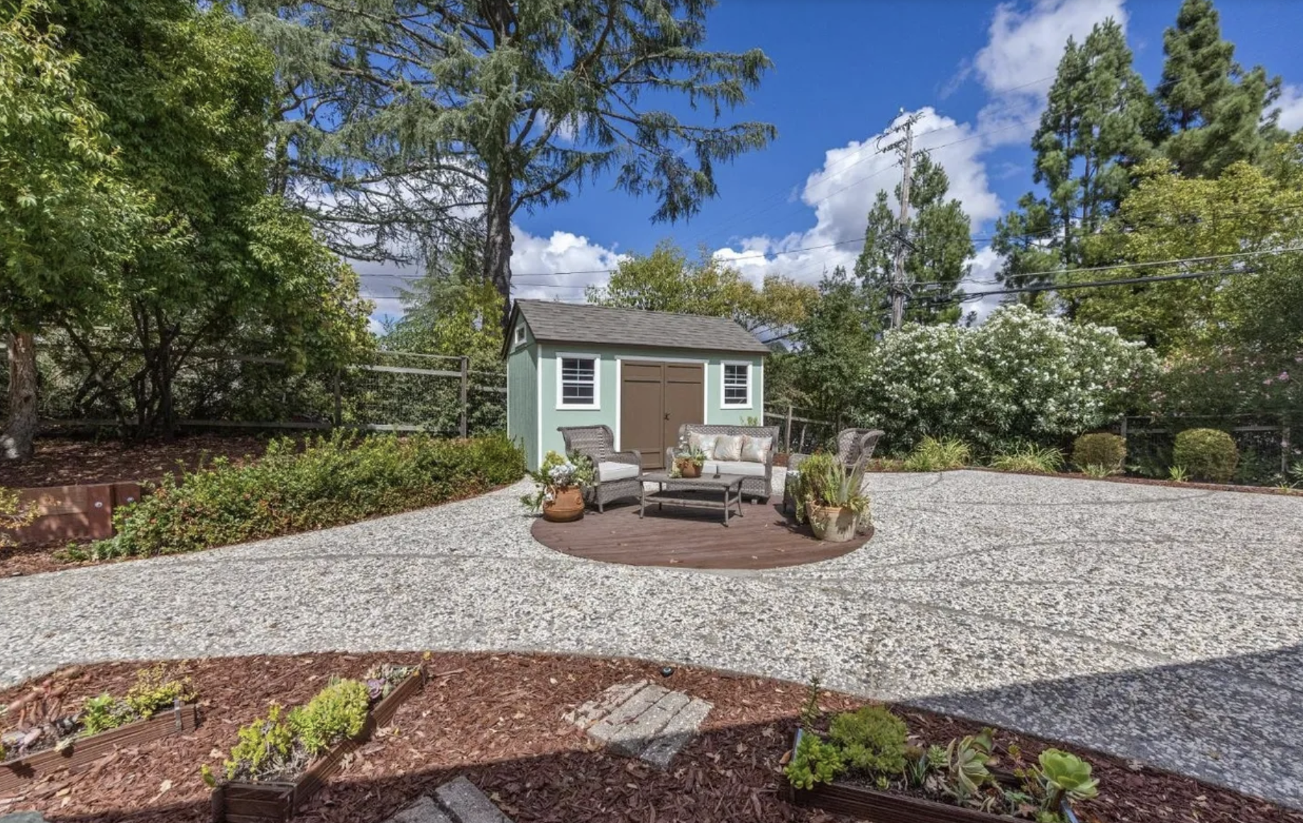 A backyard with a gravel patio, a small shed, and outdoor furniture surrounded by trees and plants.