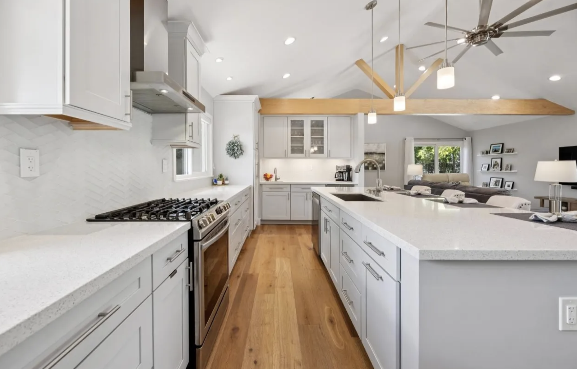 Modern open-concept kitchen with white cabinets, a large island with a sink, stainless steel stove, and wooden flooring, adjacent to a living room with shelves, windows, and ceiling fans.
