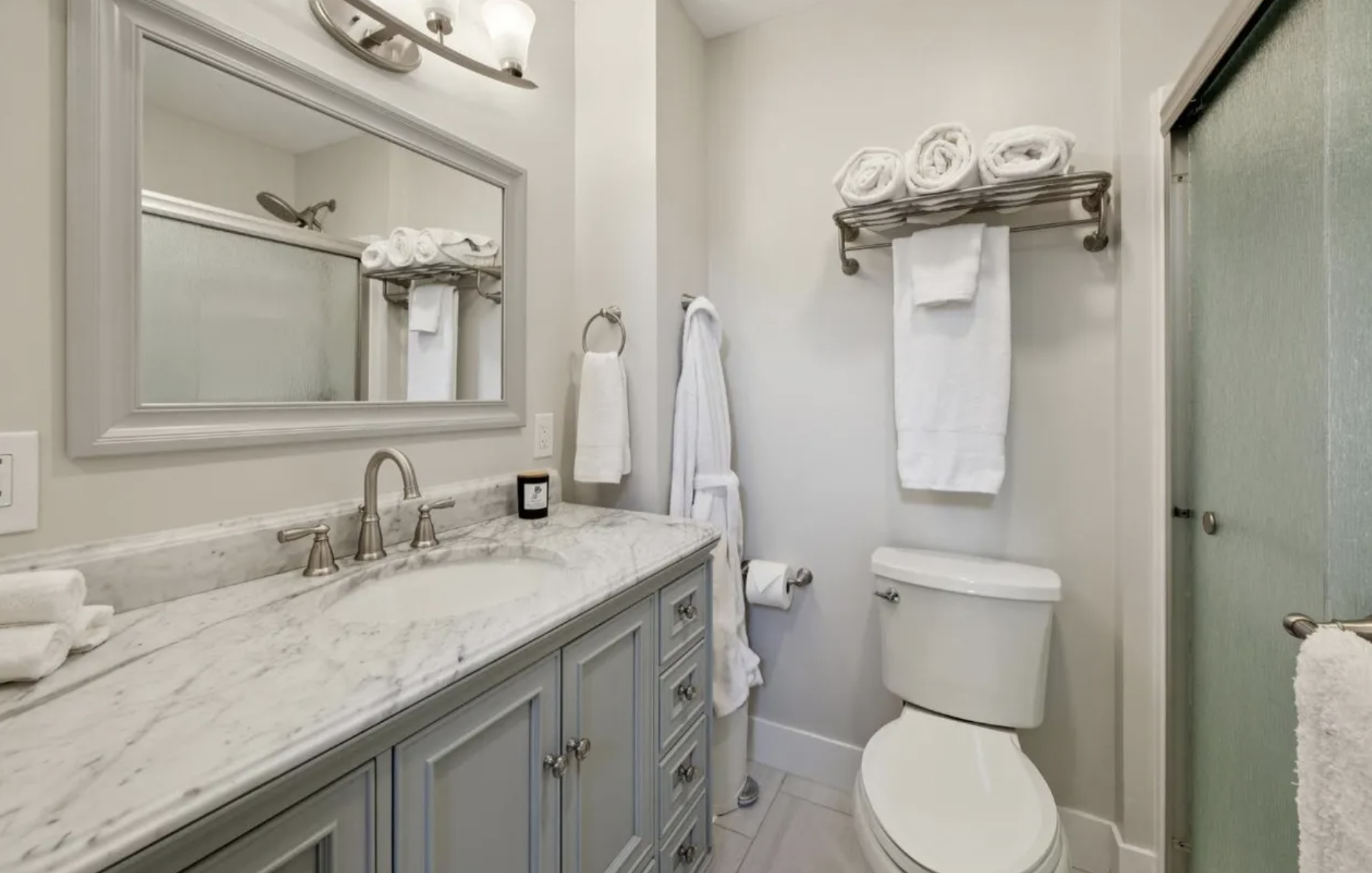Bathroom with a white marble countertop vanity, a large mirror, a faucet, neatly folded towels, a toilet, and floating shelves with rolled towels.