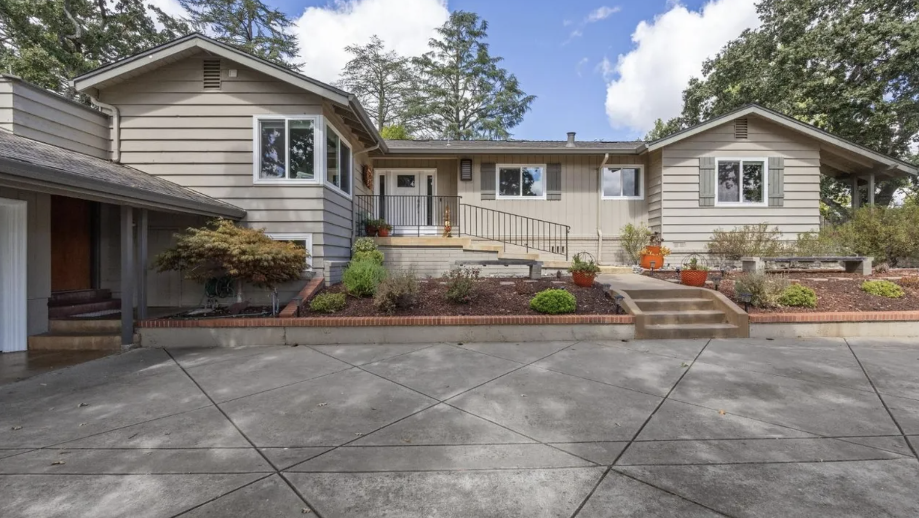 Front view of a house with a garden and stairs leading up to the entrance. The house has beige siding, multiple windows, and a small porch. The garden is landscaped with shrubs, potted plants, and mulch, and the foreground shows a concrete driveway.