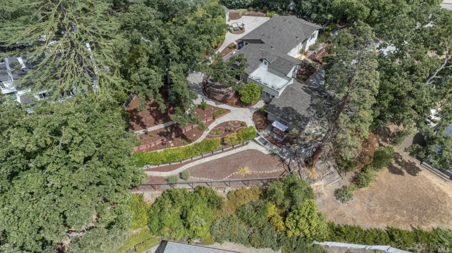 Aerial view of a residential property with a yard, trees, walkways, and multiple houses surrounded by greenery and a fence.