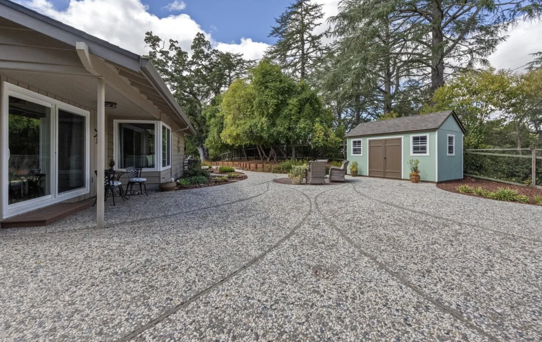 Backyard patio with a house on the left, a patio set in the middle, and a garden shed on the right, surrounded by trees and a fenced yard.