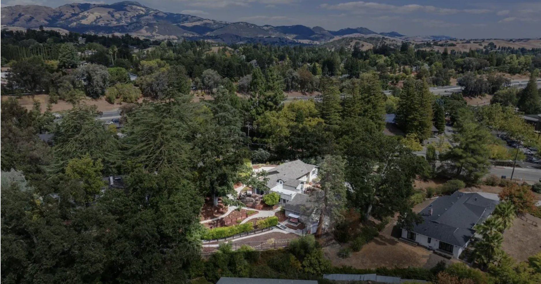 Aerial view of a neighborhood with houses surrounded by tall trees and mountains in the background.