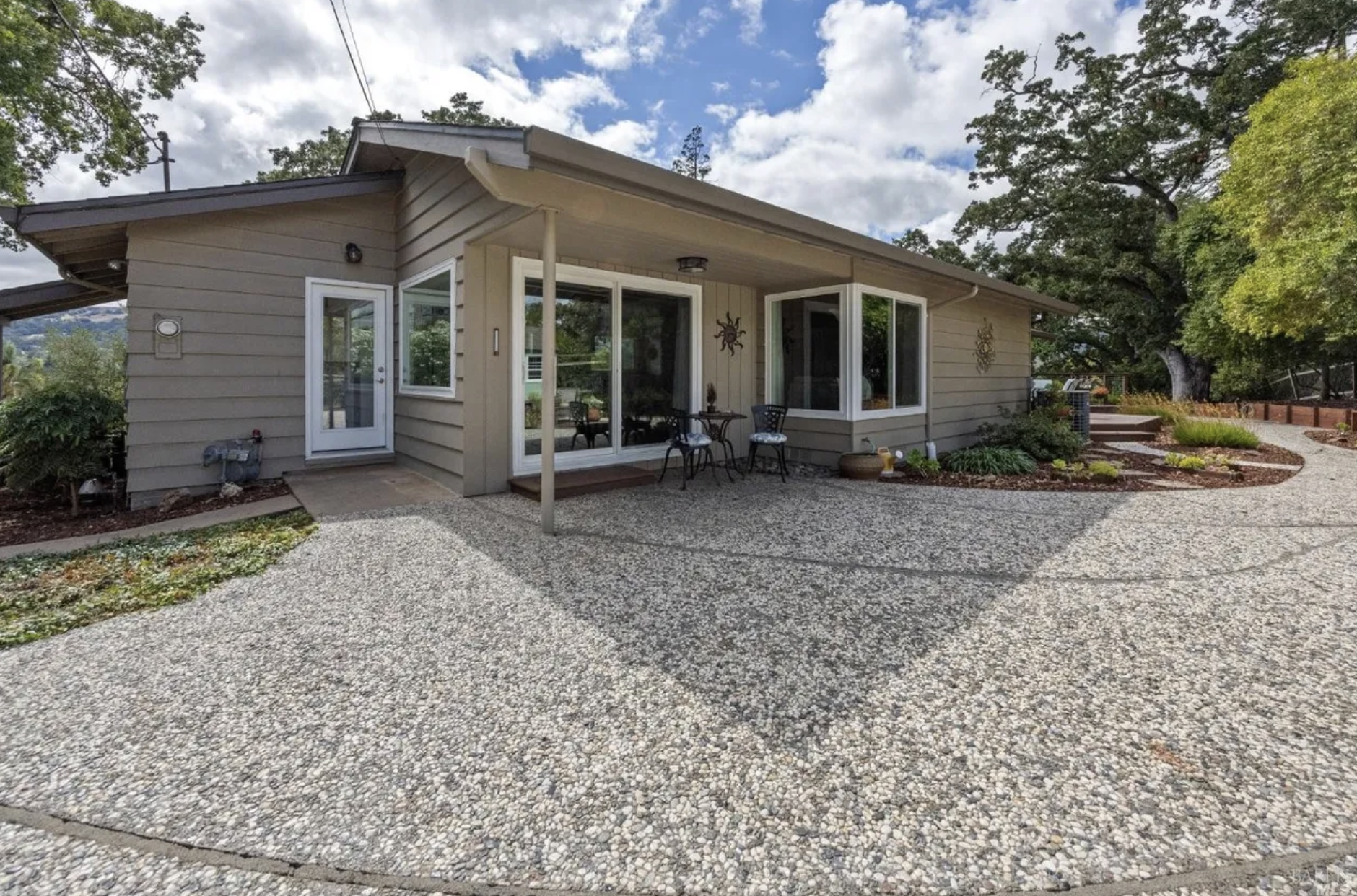 A house with beige siding and large windows, small patio with two chairs and table, gravel driveway, landscaped yard with plants and trees.