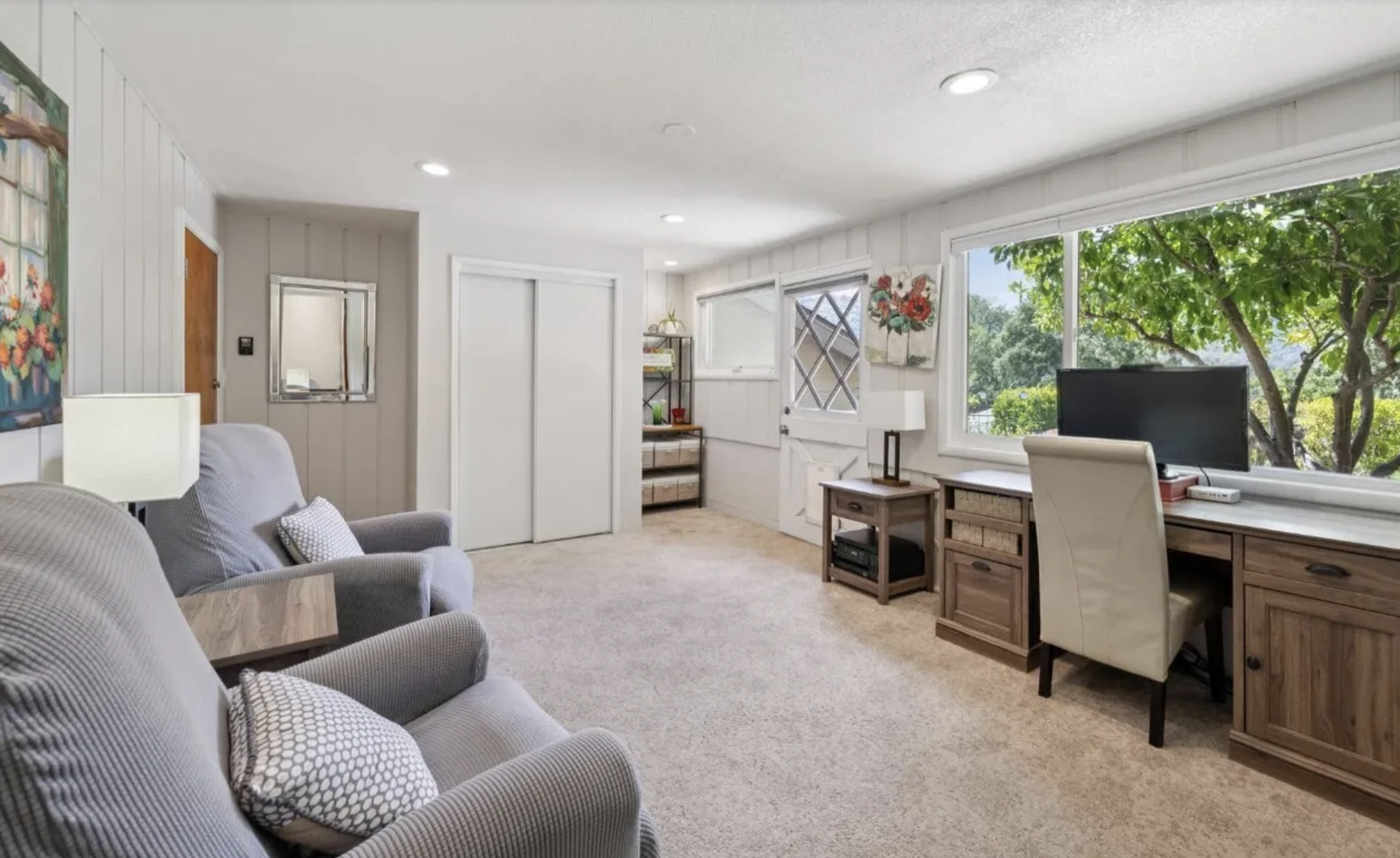 A living room with two armchairs and a sofa, a side table, a desk with a computer, large windows showing trees outside, and a door with a diamond-shaped window.