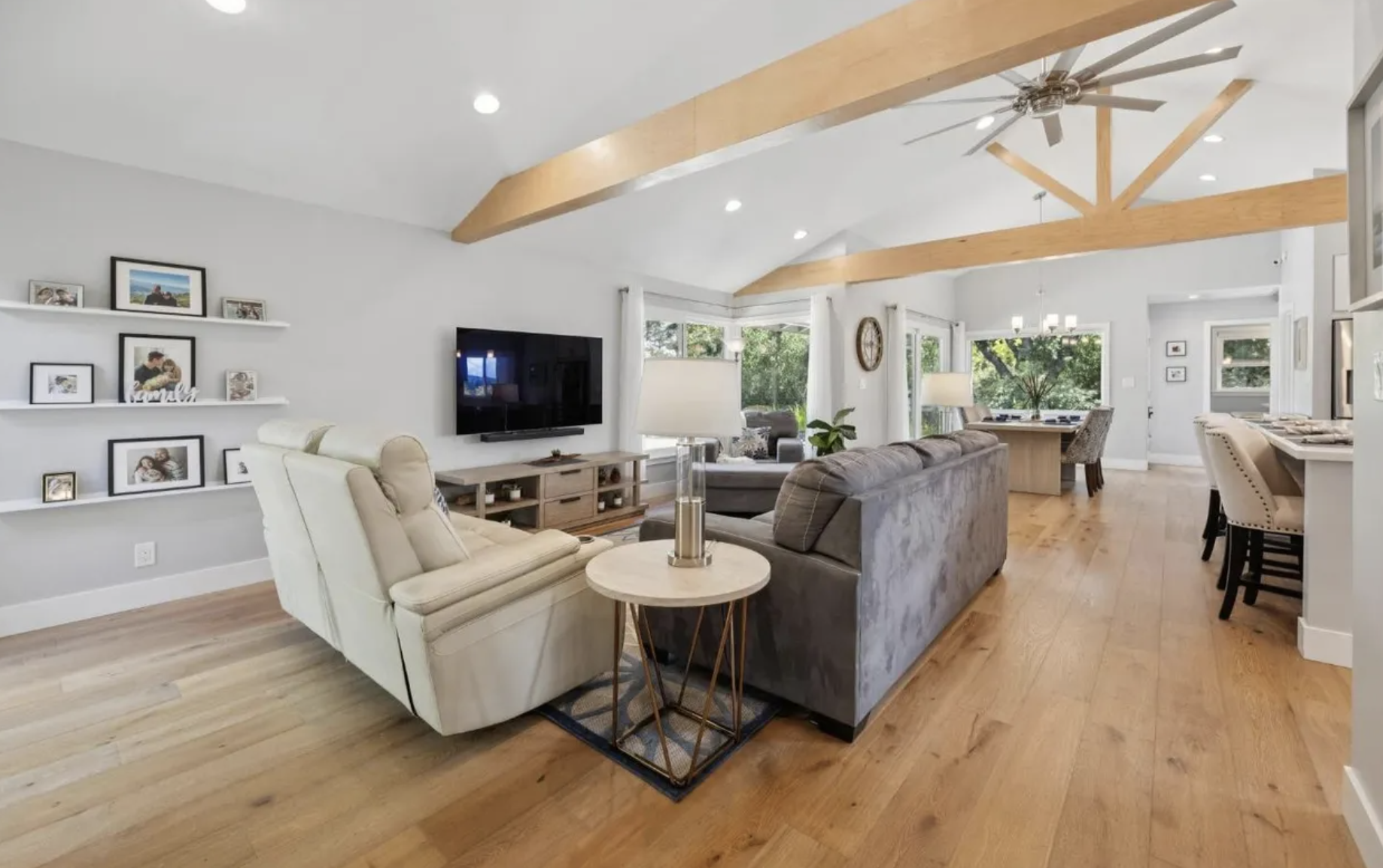 A spacious living room with wooden beams on the ceiling, a large window with white curtains, a gray sofa, a cream armchair, a flat-screen TV, and a dining area in the background with large windows and outdoor greenery visible.