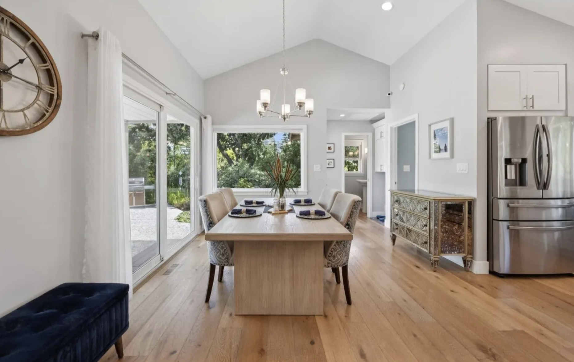 Bright dining room with a light wood table set for six, beige upholstered chairs, a modern chandelier, large windows with white curtains, a sliding glass door, and a view of a backyard with trees.