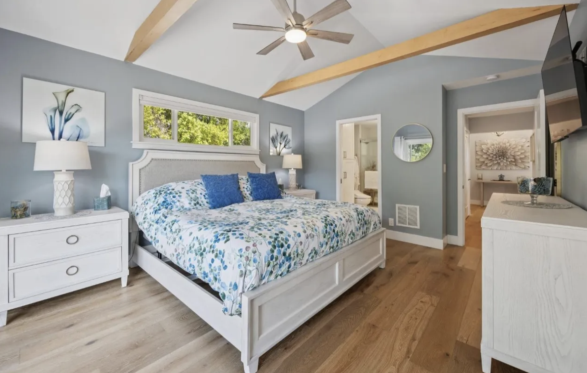 A bedroom with a large bed, white nightstands, lamps, and blue decor, featuring a ceiling fan and wood floors.
