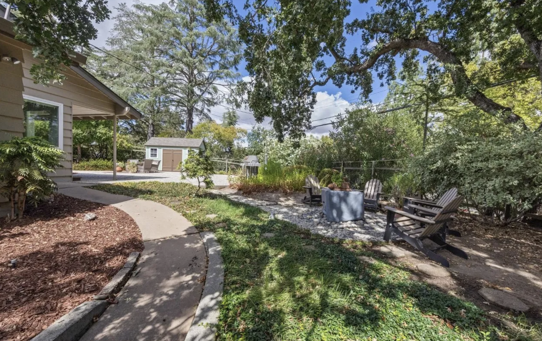 A backyard with a curved concrete pathway, a fire pit with Adirondack chairs around it, surrounded by trees and bushes, with a small shed and patio furniture in the background under a partly cloudy sky.