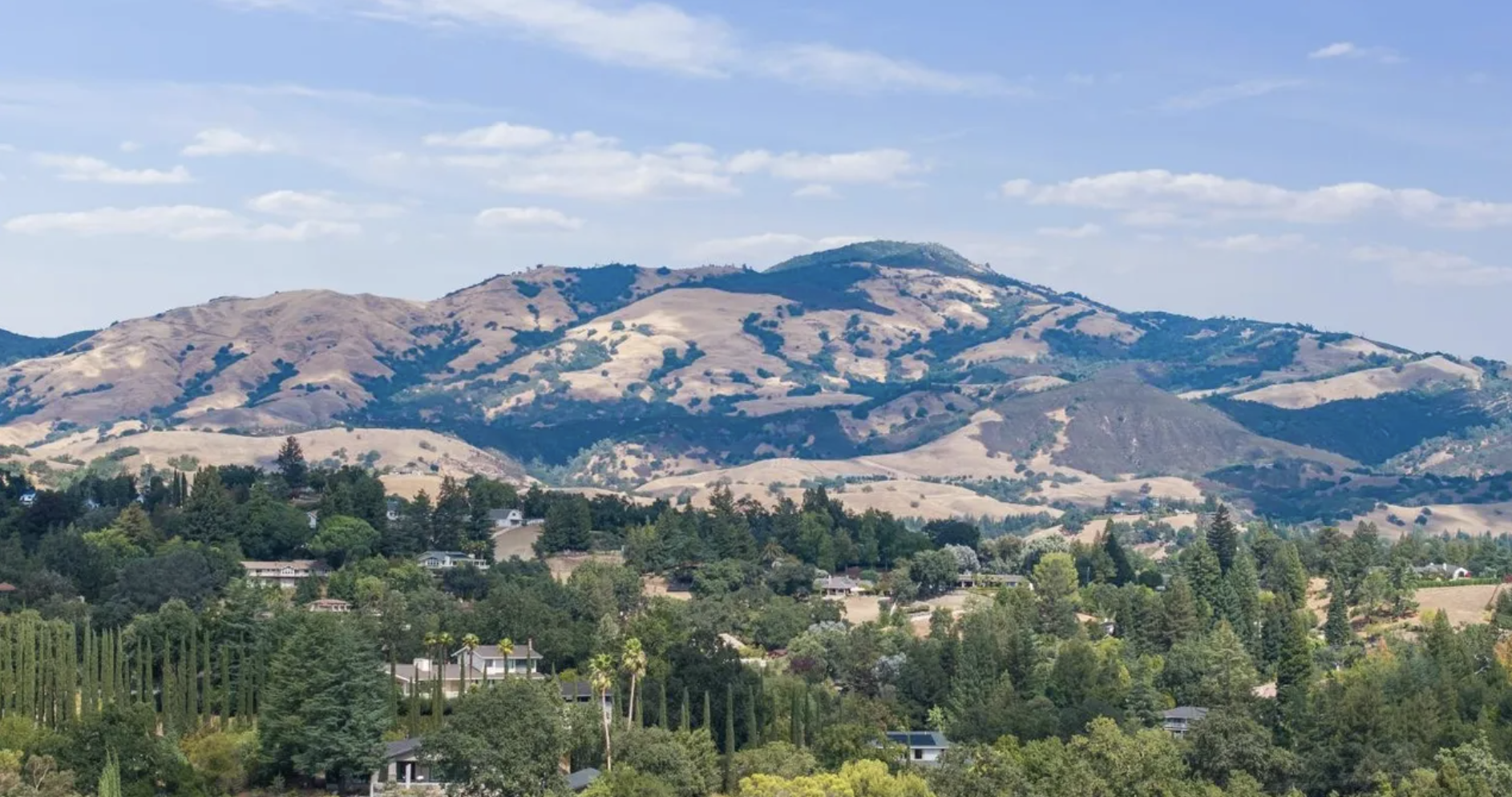 Scenic view of rolling hills with dry grass and patches of green, scattered trees, and residential houses in the foreground, under a partly cloudy blue sky.