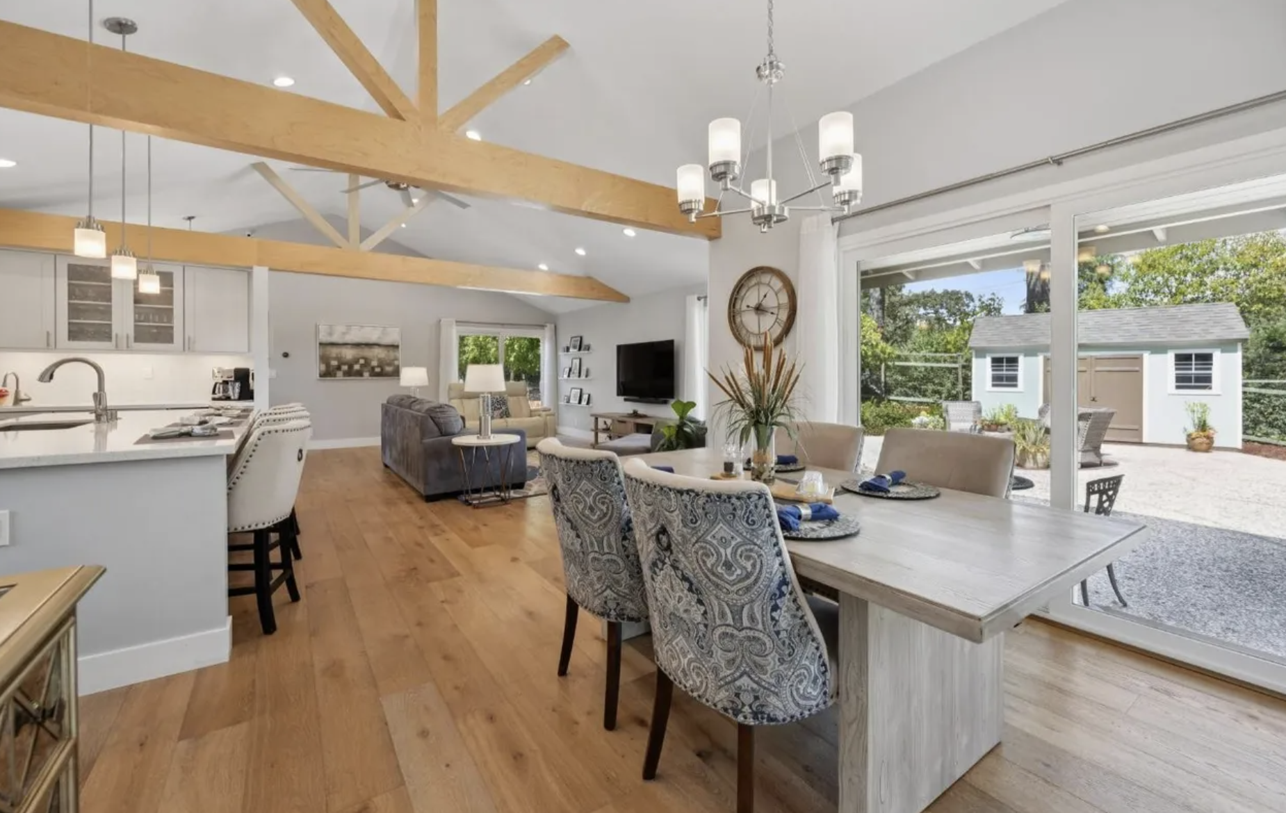 Open-concept living and dining area with hardwood floors, large sliding glass doors leading to a backyard, a wooden dining table with chairs, a gray sofa, a mounted flat-screen TV, and a kitchen area with an island and modern appliances.
