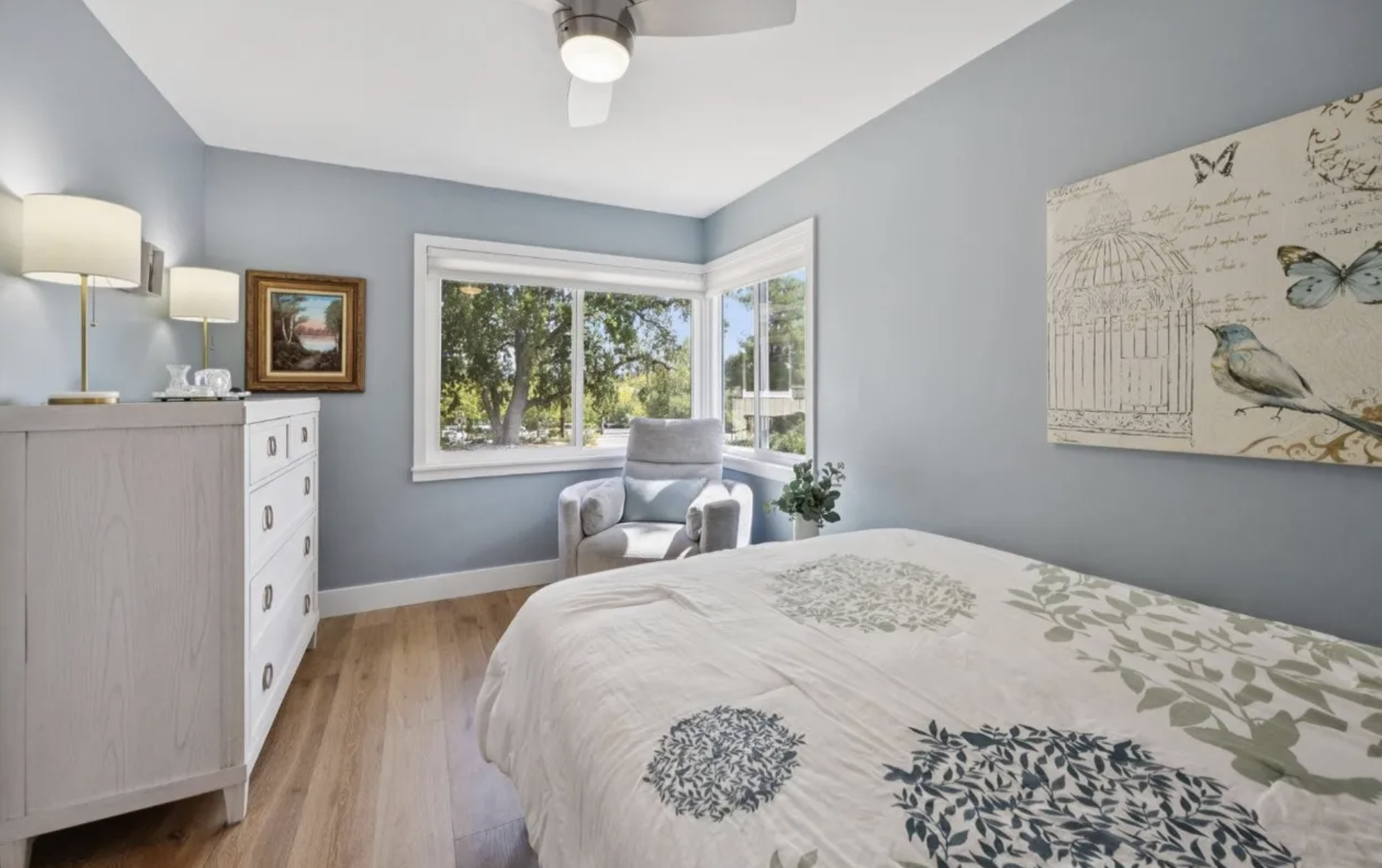 A cozy bedroom with light blue walls, a white bed with botanical patterned bedding, a window with a view of trees, a white armchair, a dresser with lamps, and wall art featuring butterflies and a birdcage.