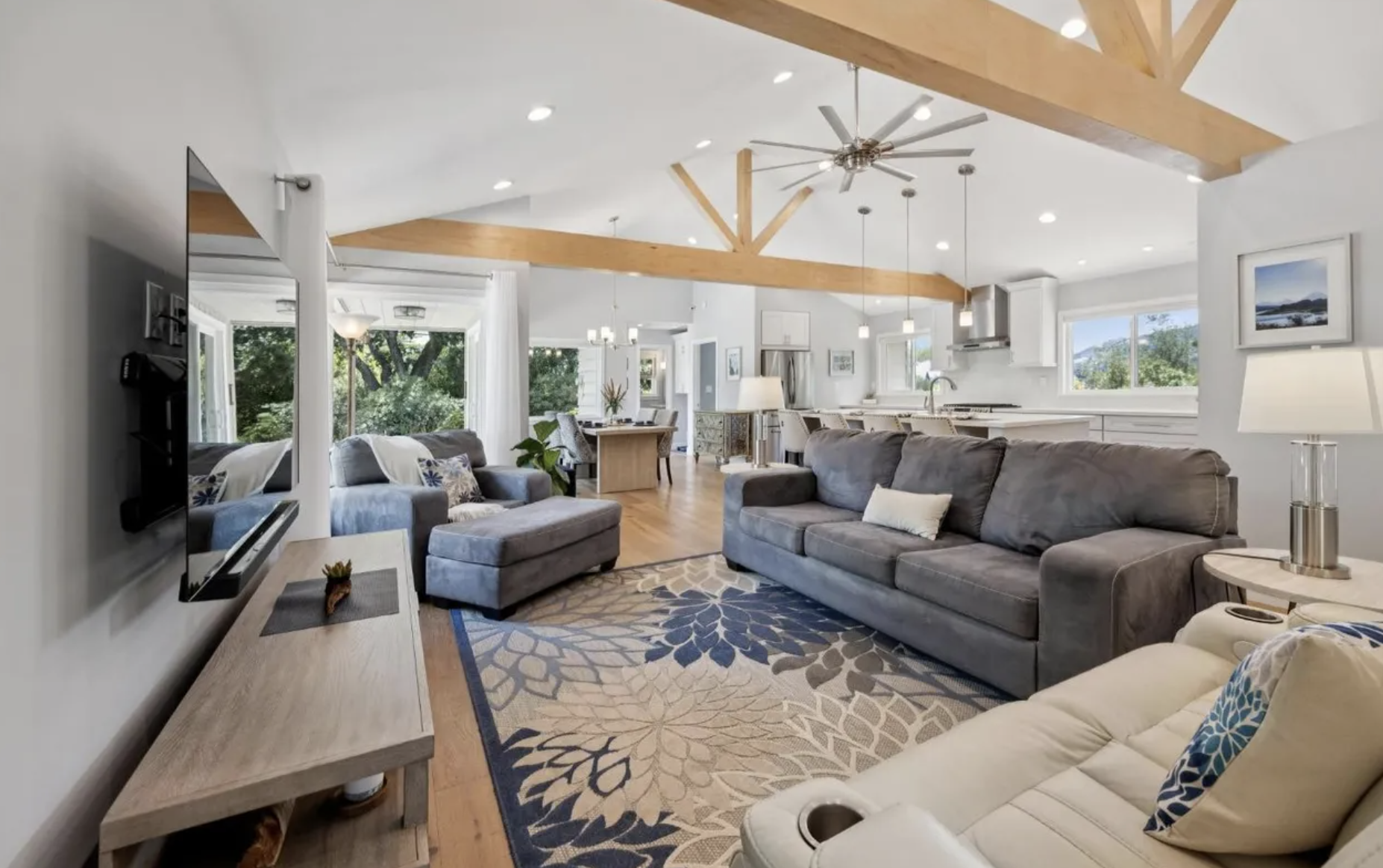 Open-concept living room with gray sofas, a cream recliner, a wood coffee table, a colorful floral area rug, large windows, and a view of the kitchen and dining area. Exposed wooden beams and a ceiling fan are also visible.