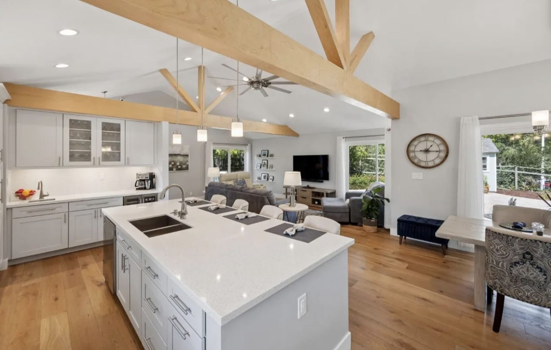 Open-concept living room and kitchen with white cabinetry, wooden beams, hardwood floors, and large windows with outside greenery.