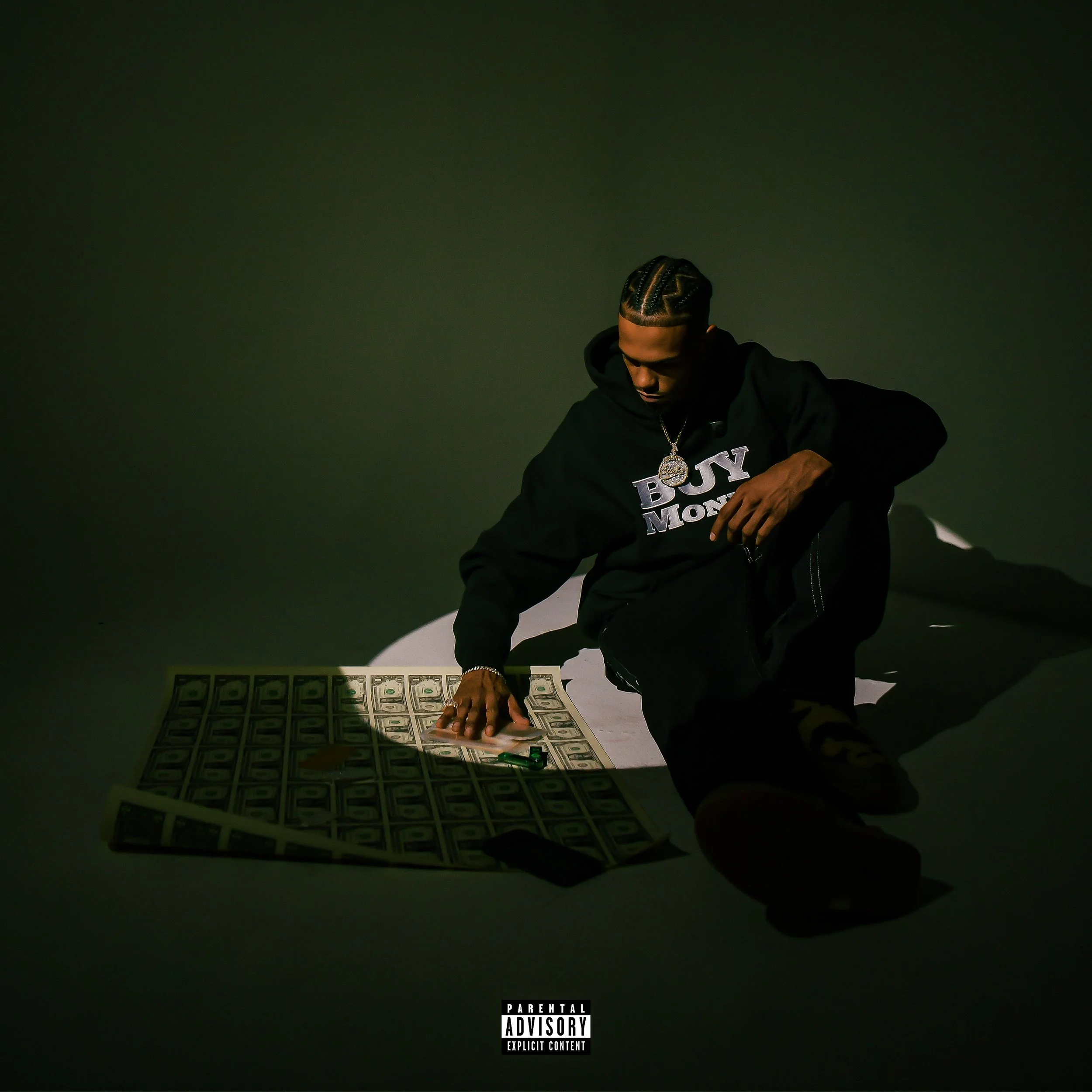 A man kneeling on the ground, surrounded by scattered US dollar bills, with a shadow cast on the floor. He is wearing a black hoodie with the words 'BOY MAMA' and a large chain with a medallion.